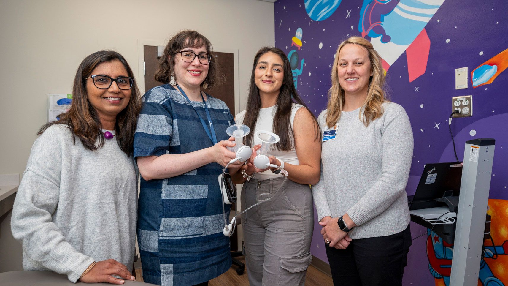Front left to right, Dr. Dharod, Dr. DeJesus, grad student Salena Villa, and Julie Wenzel standing in a room with colorful space-themed wall art, holding breast pump equipment and tubing, with a laptop on a nearby table.