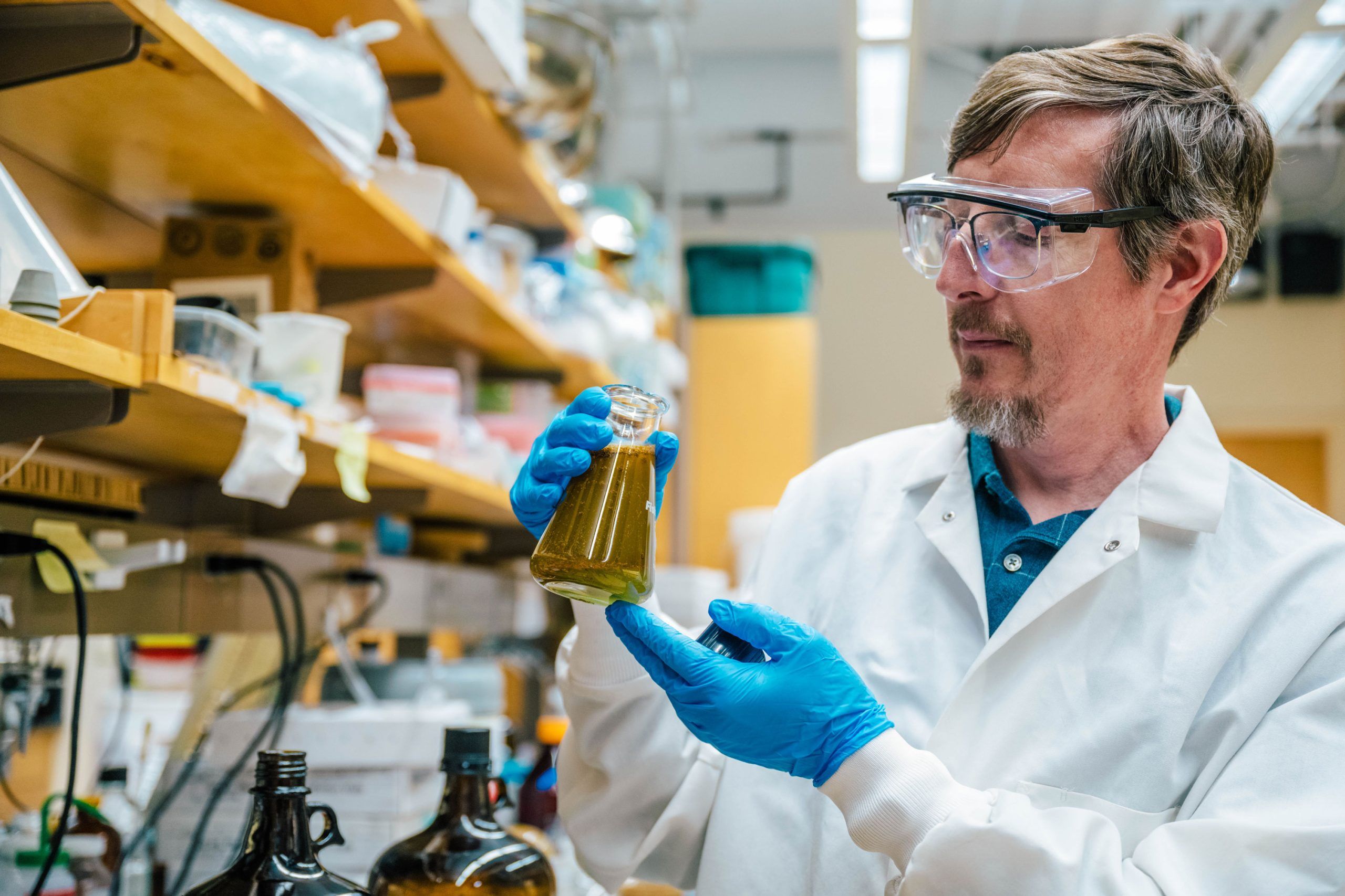 Tyler Graf wearing a white lab coat and blue gloves holding a glass flask filled with greenish liquid in a laboratory. Shelves with bottles, containers, and equipment are visible in the background.