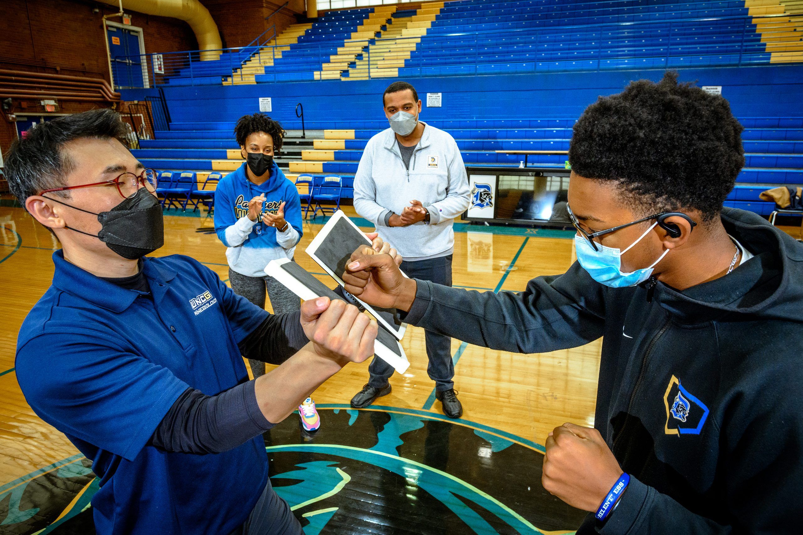 A high school student hits a punch pad held by Yongsun Lee, while Dr. Hemphill and the school's coach look on in the background.