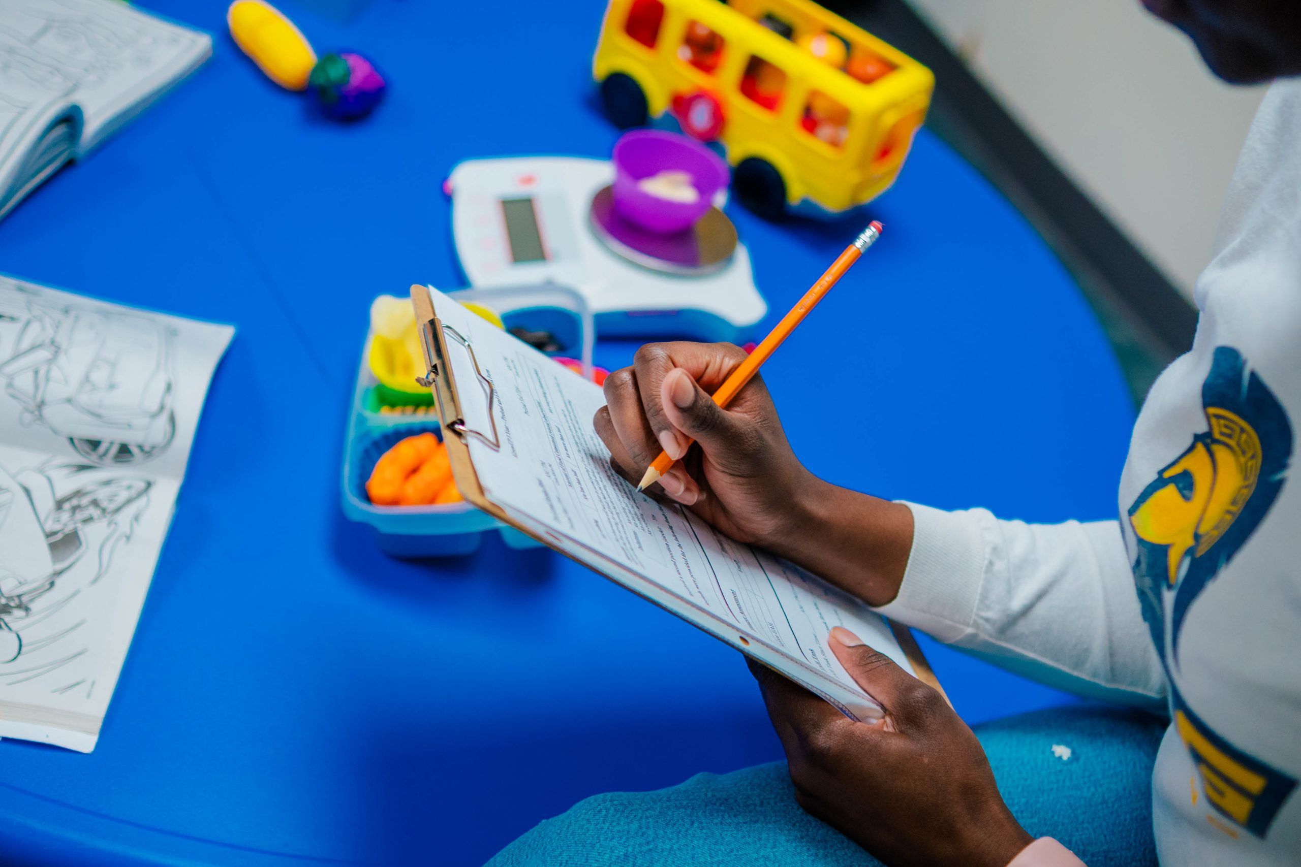 Close-up of Jahleen Gourdine writing on a clipboard with a pencil at a small blue table; the table holds open workbooks, colorful educational toys, and a yellow toy bus.
