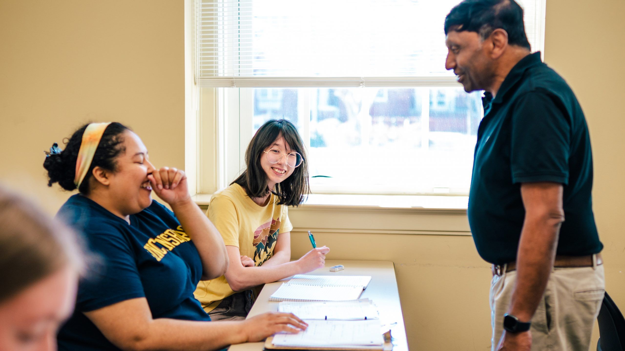 Dr. Shivaji standing near a desk speaking with two students who are seated and writing in notebooks. One of the students is smiling while the other is laughing.