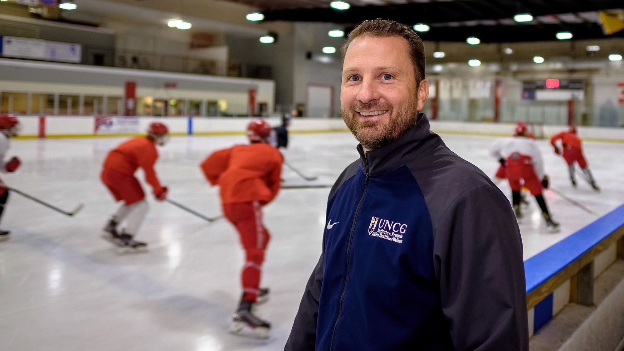 Dr. Milroy standing at an ice rink with a youth hockey game in the background.