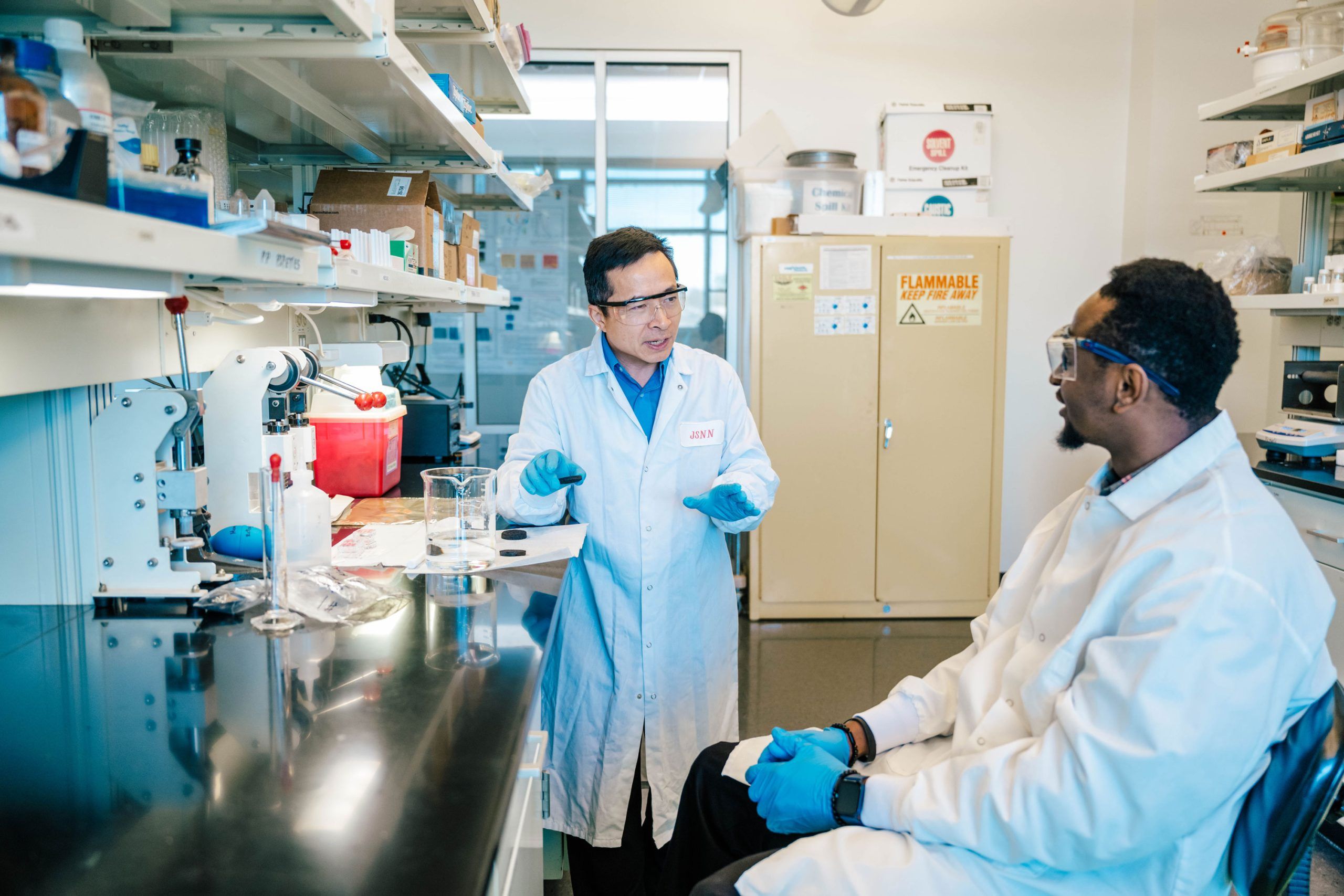 Dr. Wei and his student in a laboratory wearing white coats and blue gloves. One is standing and gesturing with hands while the other sits on a stool listening. A black countertop holds glass beakers and lab tools, and shelves above contain boxes and containers. A yellow safety cabinet labeled “Flammable Keep Fire Away” is visible in the background.