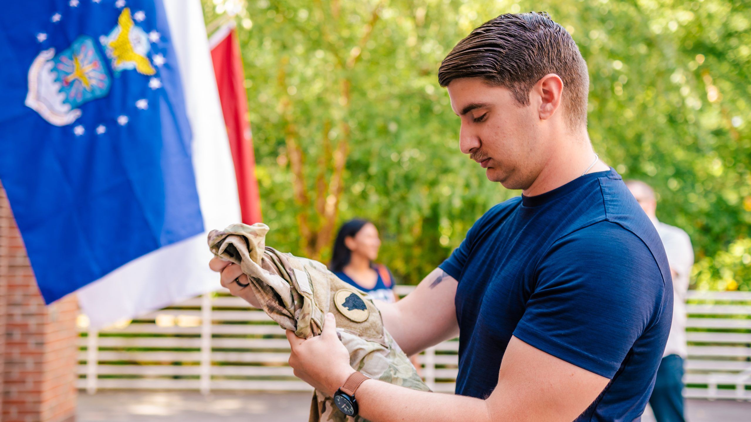 Garett Barto holding a camouflage military uniform outdoors near a flag display, with trees and a white railing in the background.