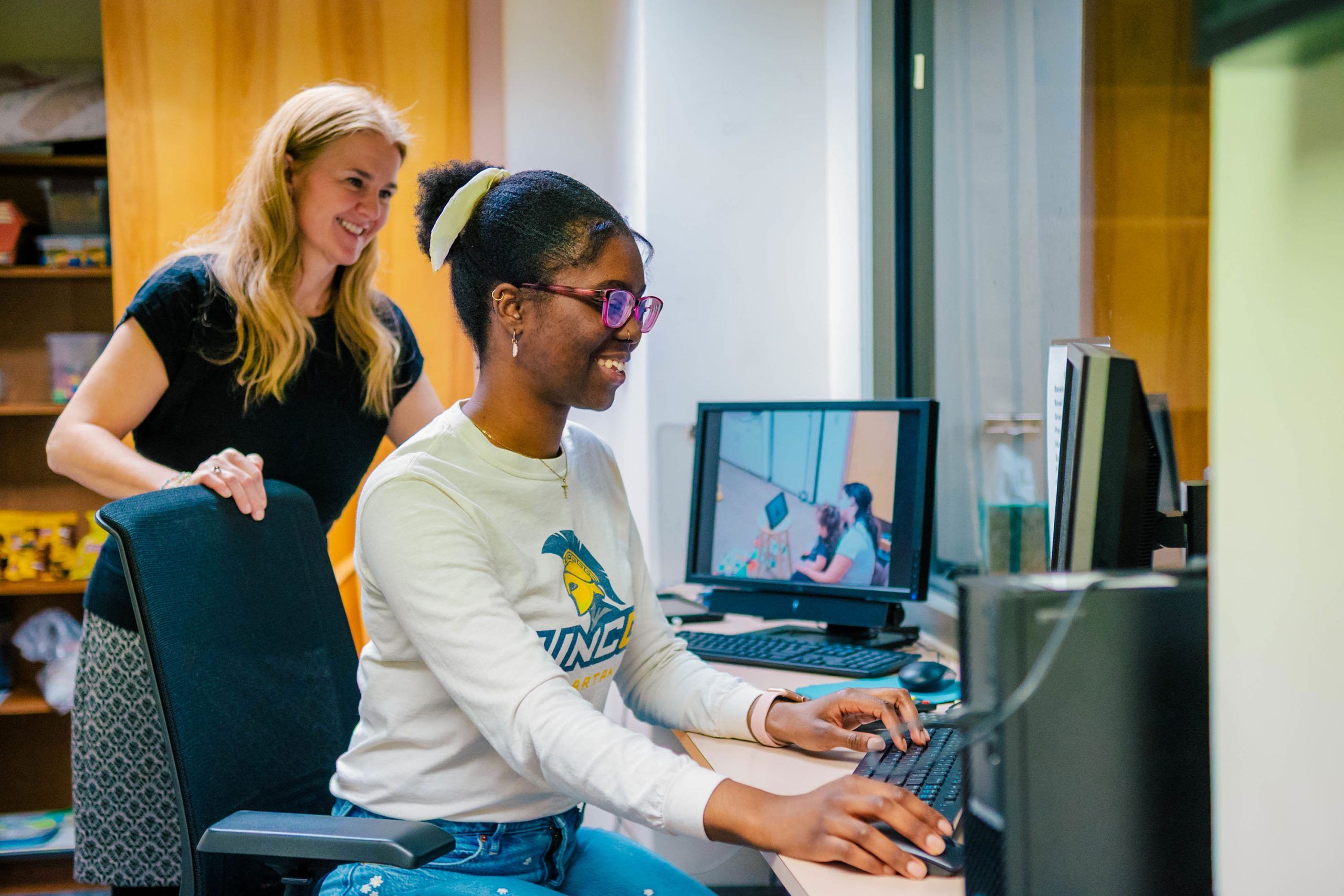 Dr. Shriver and Jahleen Gourdine working at a computer station; Gourdine seated typing on a keyboard, Dr. Shriver standing nearby. A monitor displays a video feed of the same workspace.