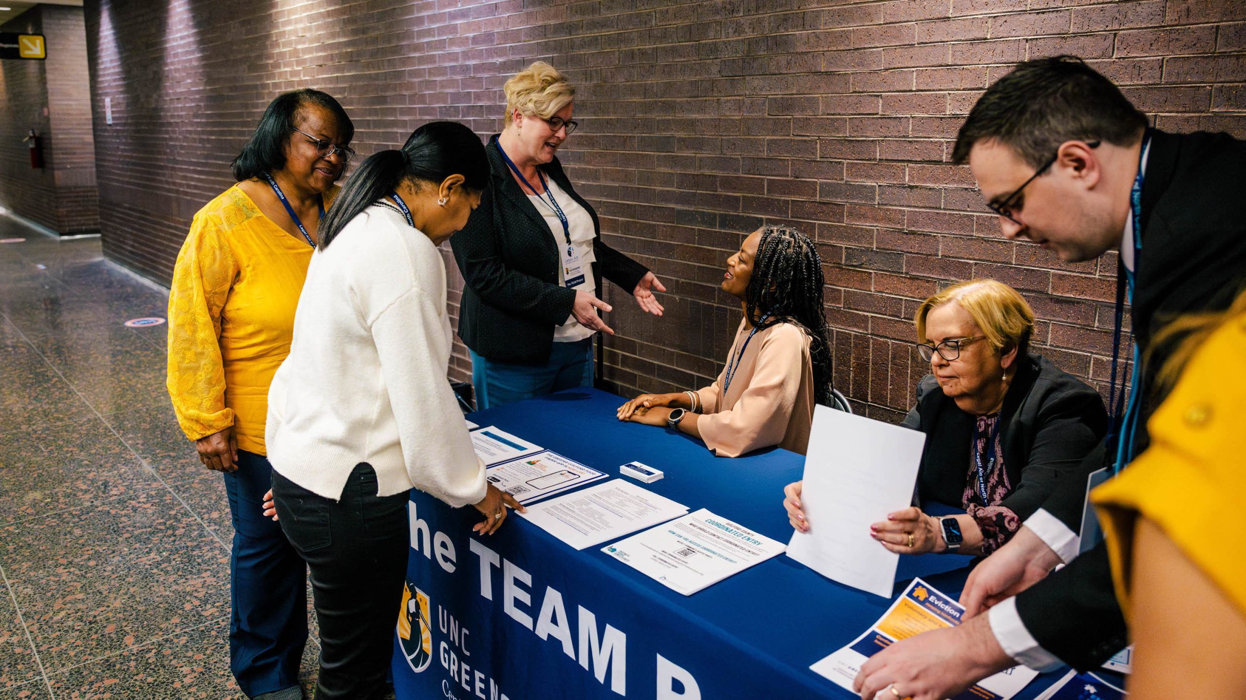 Group of people gathered at an information table with UNC Greensboro branding, reviewing documents and event materials in a hallway.