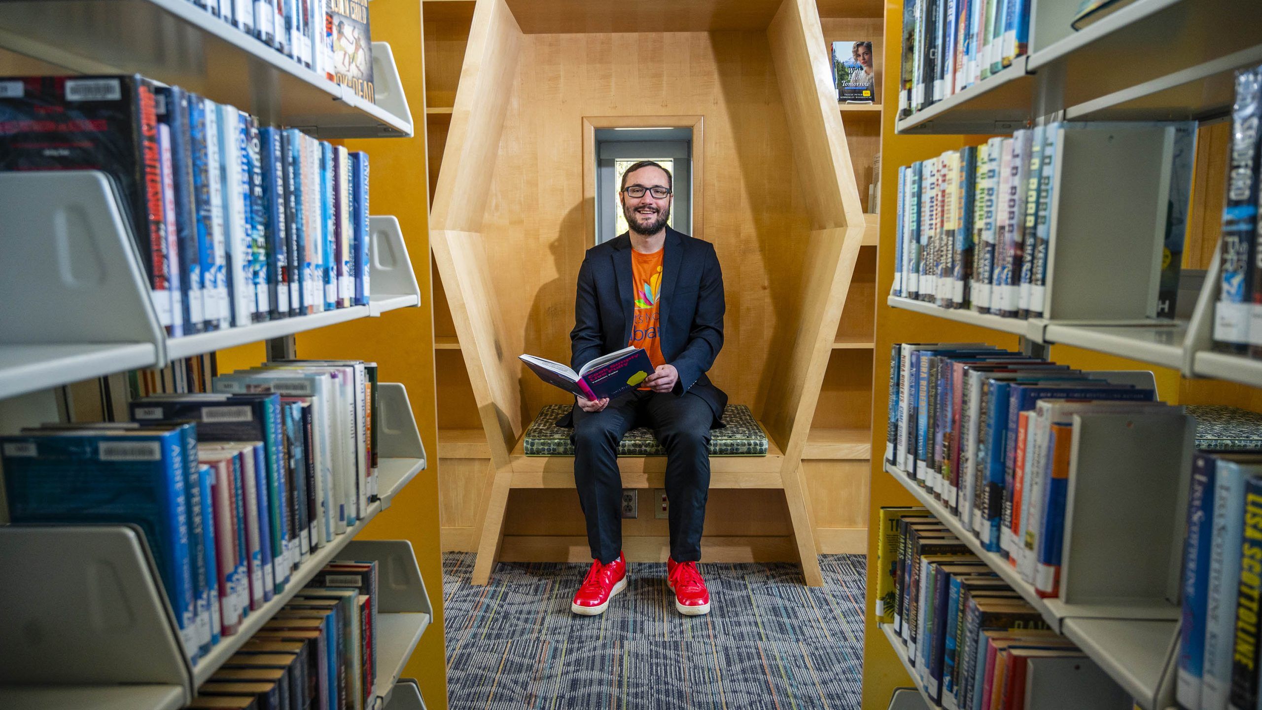 Dr. Lenstra seated in a modern wooden reading nook between bookshelves, holding an open book in a library setting.
