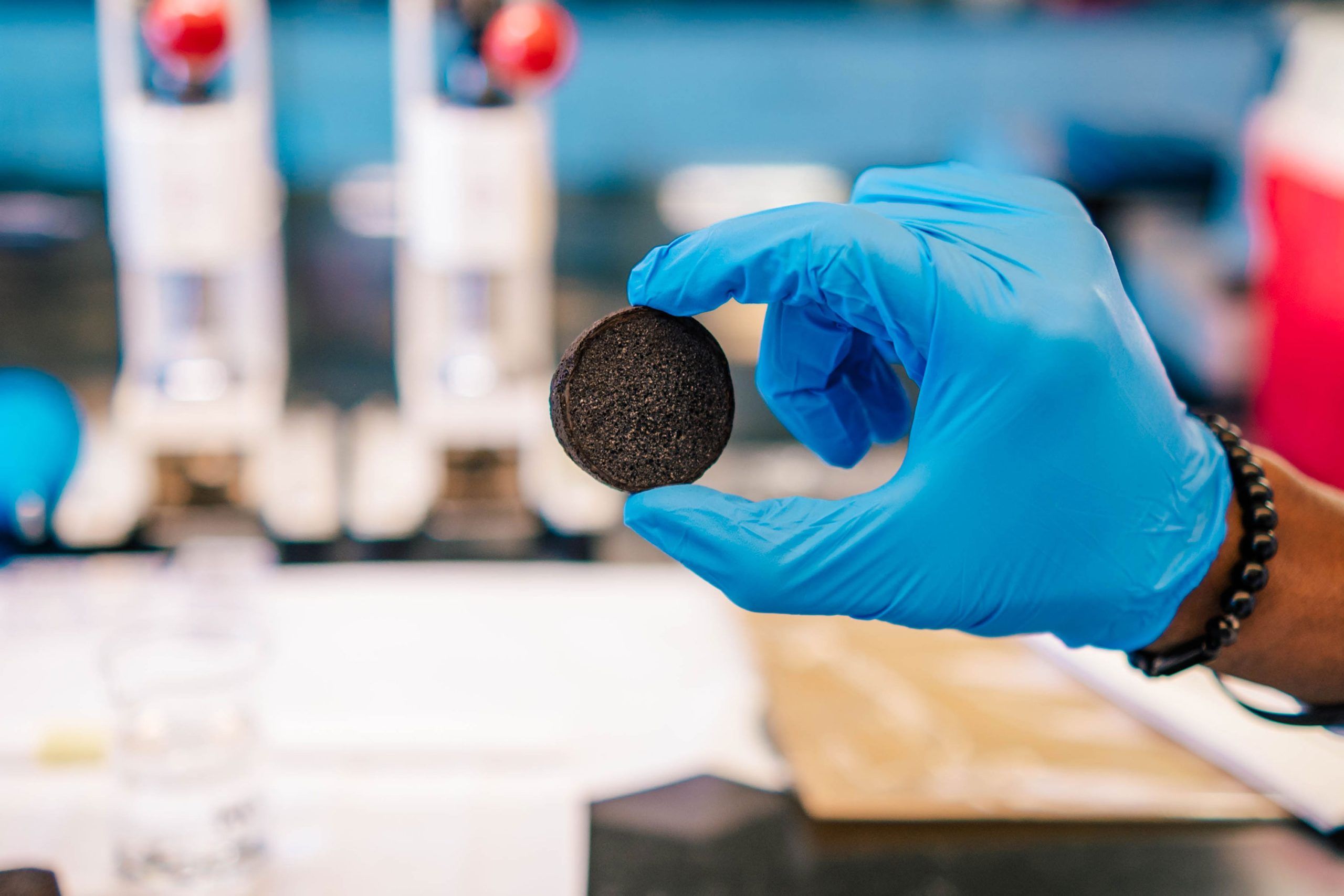 Gloved hand holding a biochar sample in a laboratory setting. The background shows blurred lab equipment and a workbench with papers and containers.