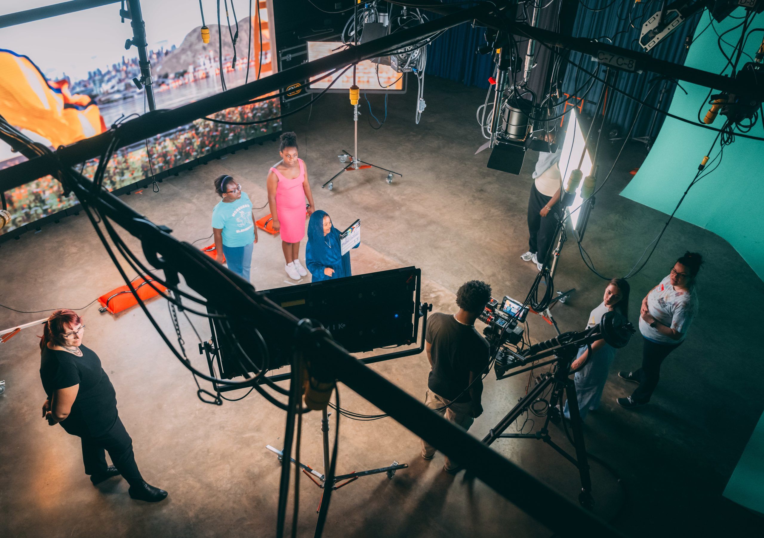 A film production studio setup with multiple people standing around a central table, preparing for a shoot. The scene includes professional video cameras on tripods, large studio lights, and overhead rigging with cables and equipment. In the background, there is a large screen displaying an image with orange panels and a green backdrop on one side, suggesting a mixed-use space for filming and virtual production. The floor has sandbags for stabilizing equipment, and the overall environment is well-lit with a controlled studio atmosphere