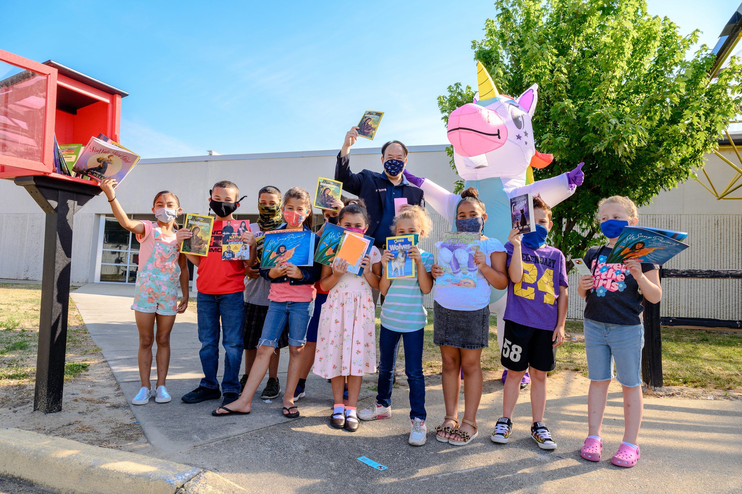 Chow, children, and the unicorn mascot holding books next to an open Little Library