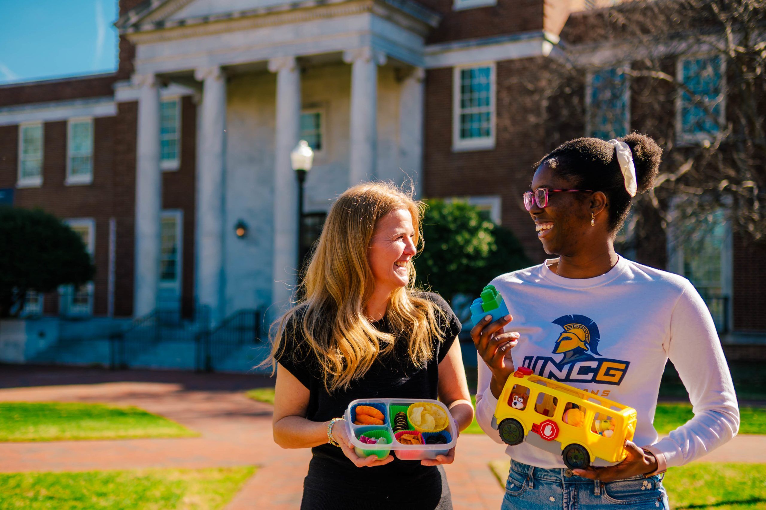 Dr. Shriver and Jahleen Gourdine standing outside a brick building with white columns, holding colorful toy food items and a yellow toy bus.