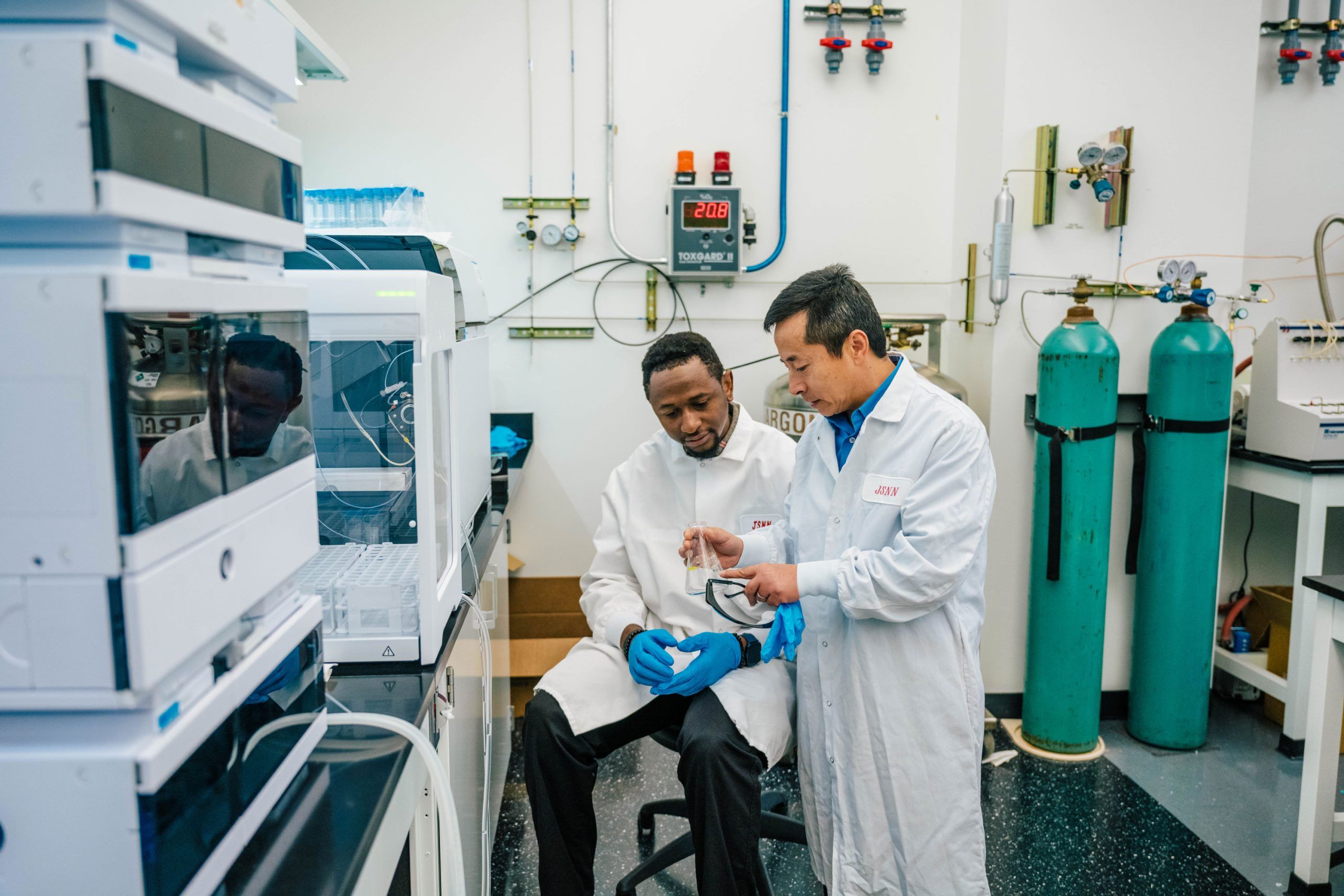 Dr. Wei and his student in a laboratory wearing white coats and blue gloves, working near a chromatography system. The student is seated while Dr. Wei points to a small glass component. The background includes gas cylinders, tubing, and a wall-mounted control panel with a digital display reading “20.8.”