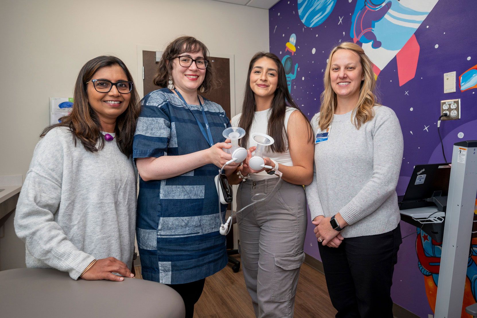 Dharod, DeJesus, grad student Selene Villa, and Wenzel standing in a colorful room with space-themed wall art, holding breast pump equipment. A computer station is visible on the right.