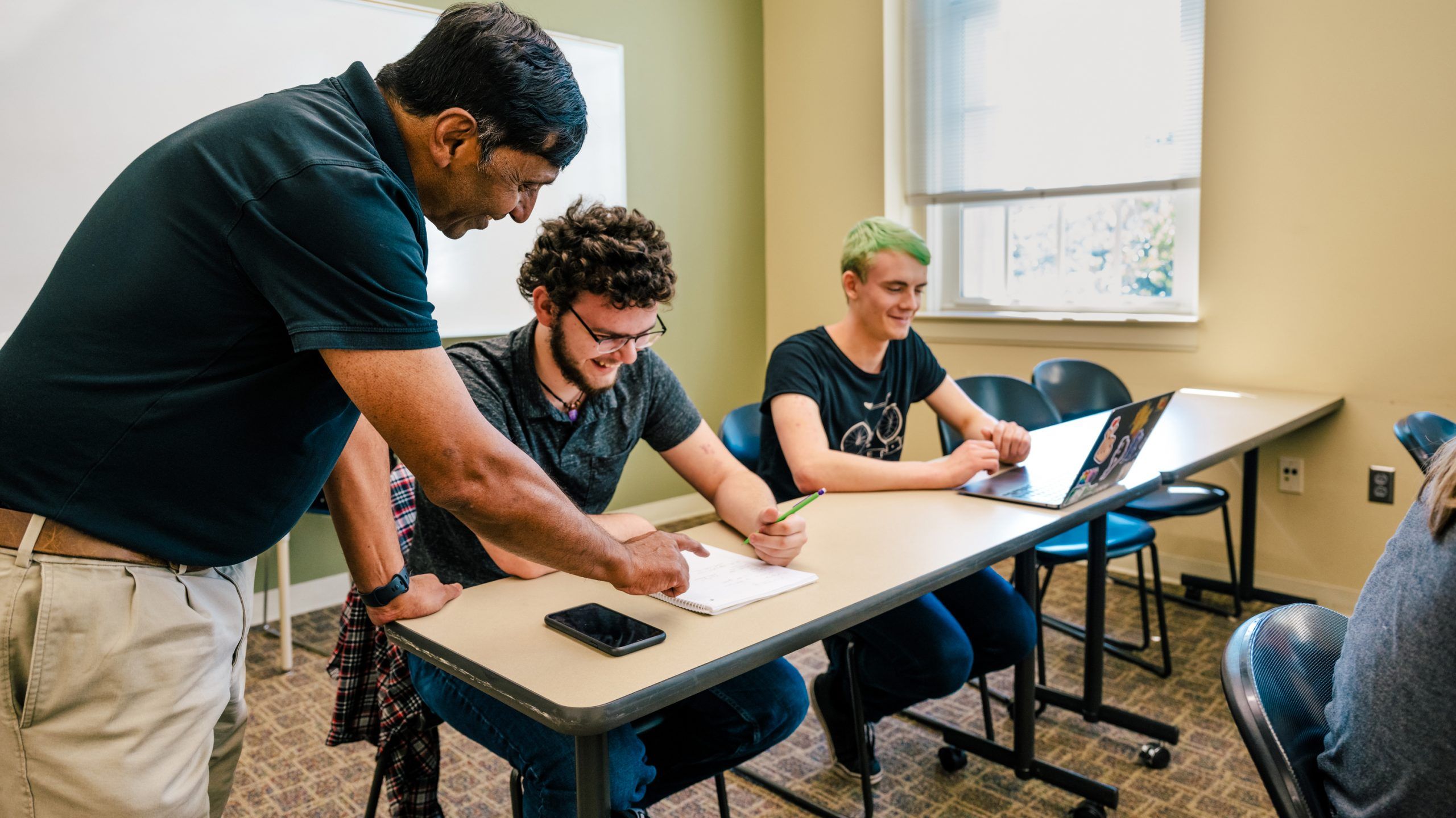 Dr. Shivaji leans over a classroom desk, pointing to a student’s notebook while discussing an assignment as another student listens and smiles.