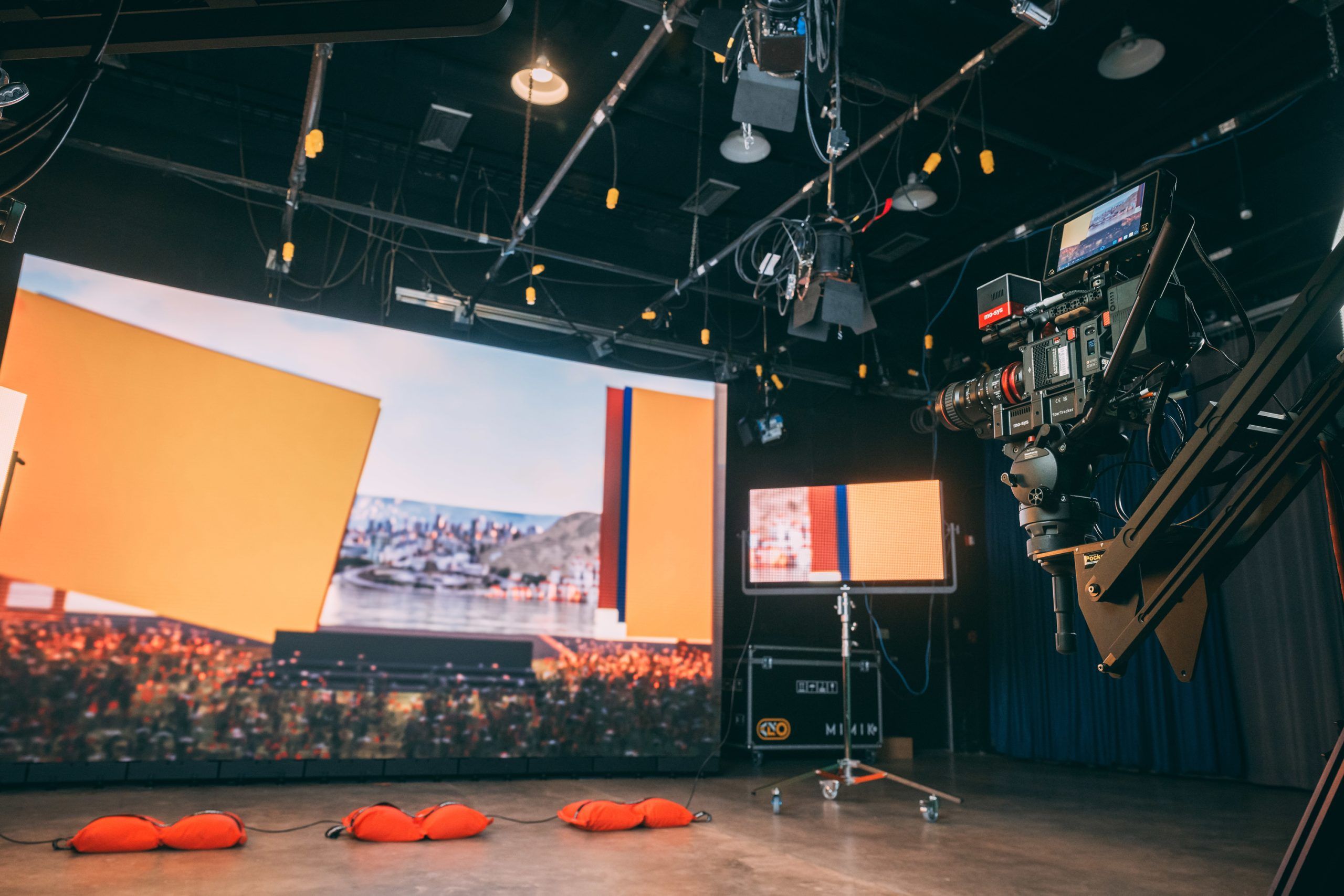 A professional video production studio with a large camera mounted on a rig in the foreground, aimed at a wide screen displaying an image of a cityscape and orange panels. The setup includes multiple studio lights hanging from the ceiling, a monitor on a stand, and several orange sandbags on the floor for equipment stabilization. The environment is dimly lit with overhead lights and cables visible, creating a controlled filming space.