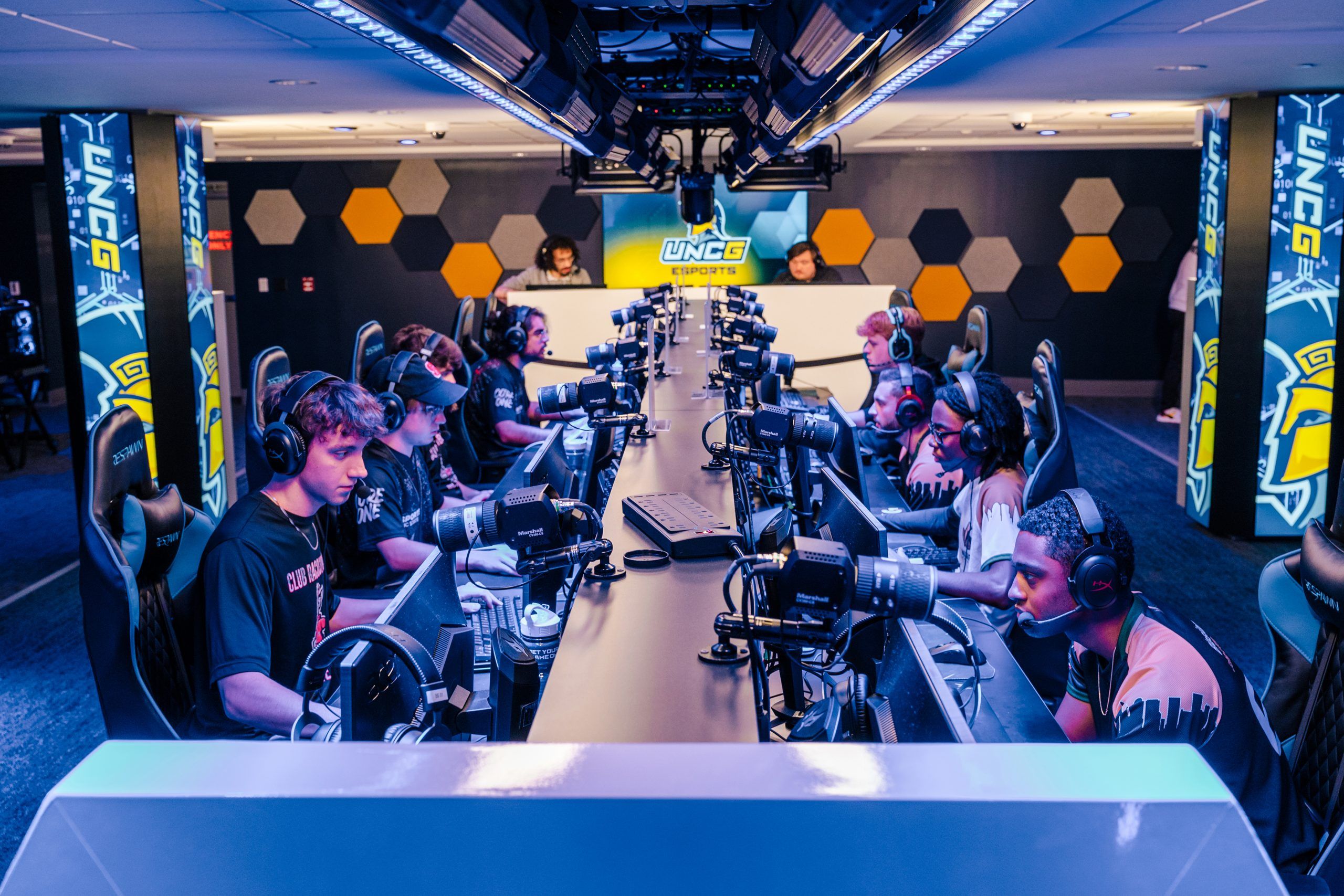 Esports players seated at a long row of gaming stations with monitors, keyboards, and headsets in a dedicated gaming arena featuring UNCG branding and hexagonal wall panels.