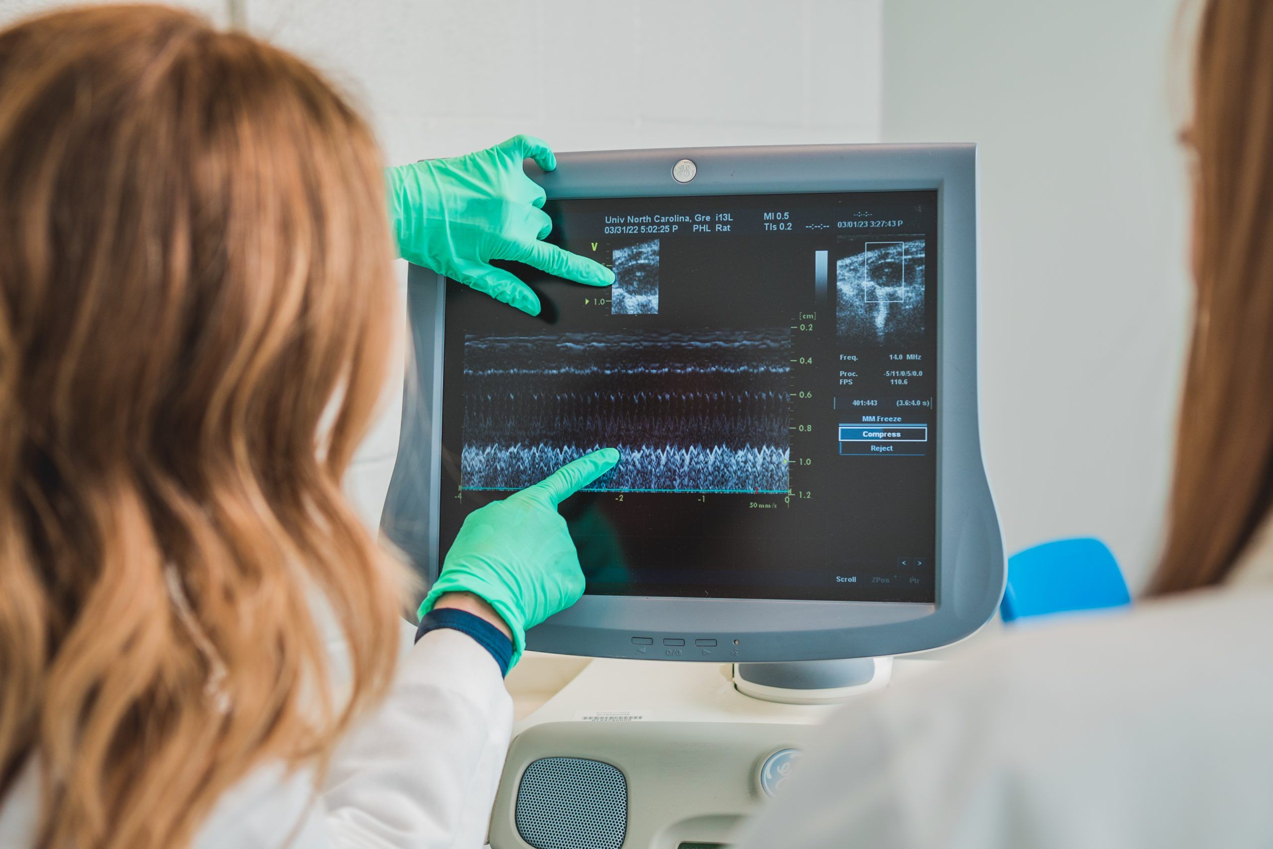 Two people in lab coats, one wearing protective gloves while pointing at a screen.