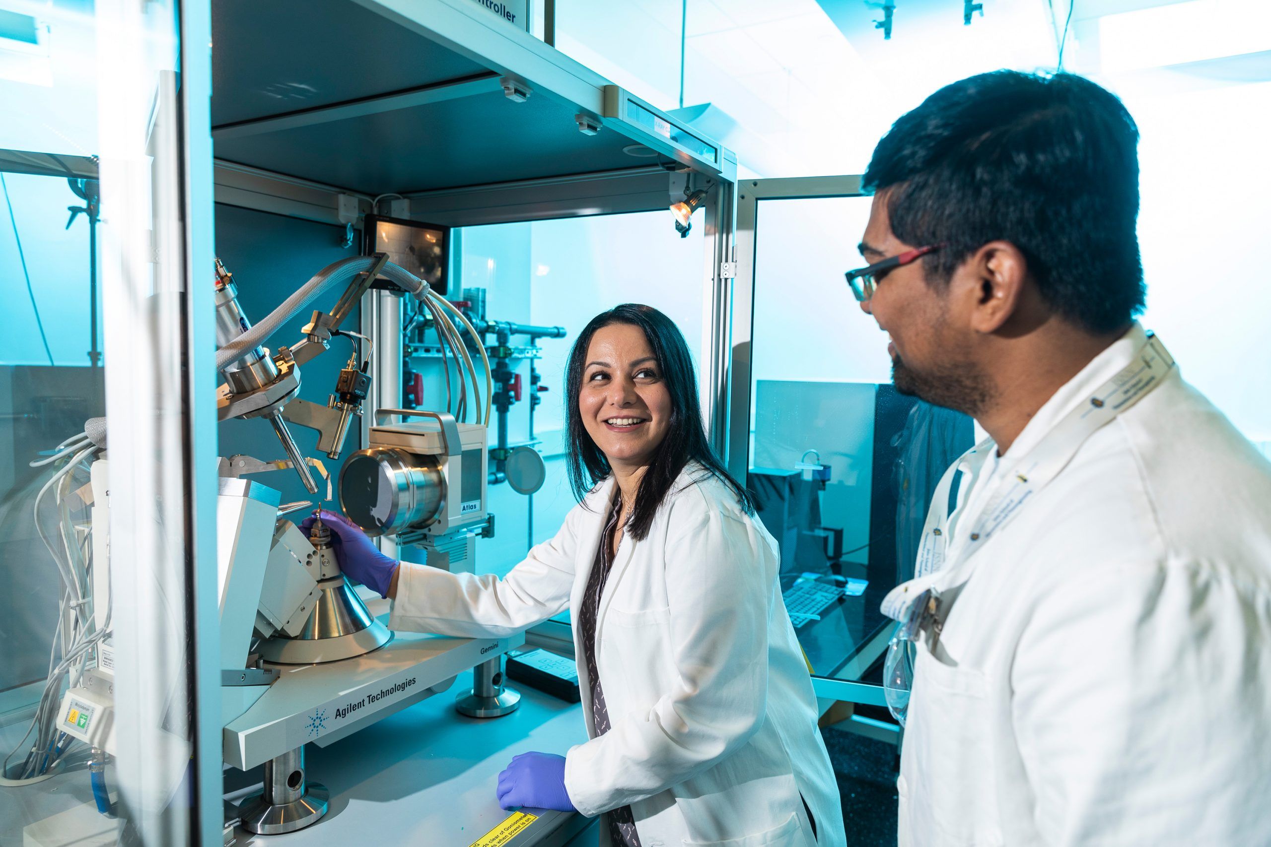 Dr. Hermatian wearing a lab coat while loading a sample into the x-ray crystallographer as she smiles at postdoc Firoz Khan.