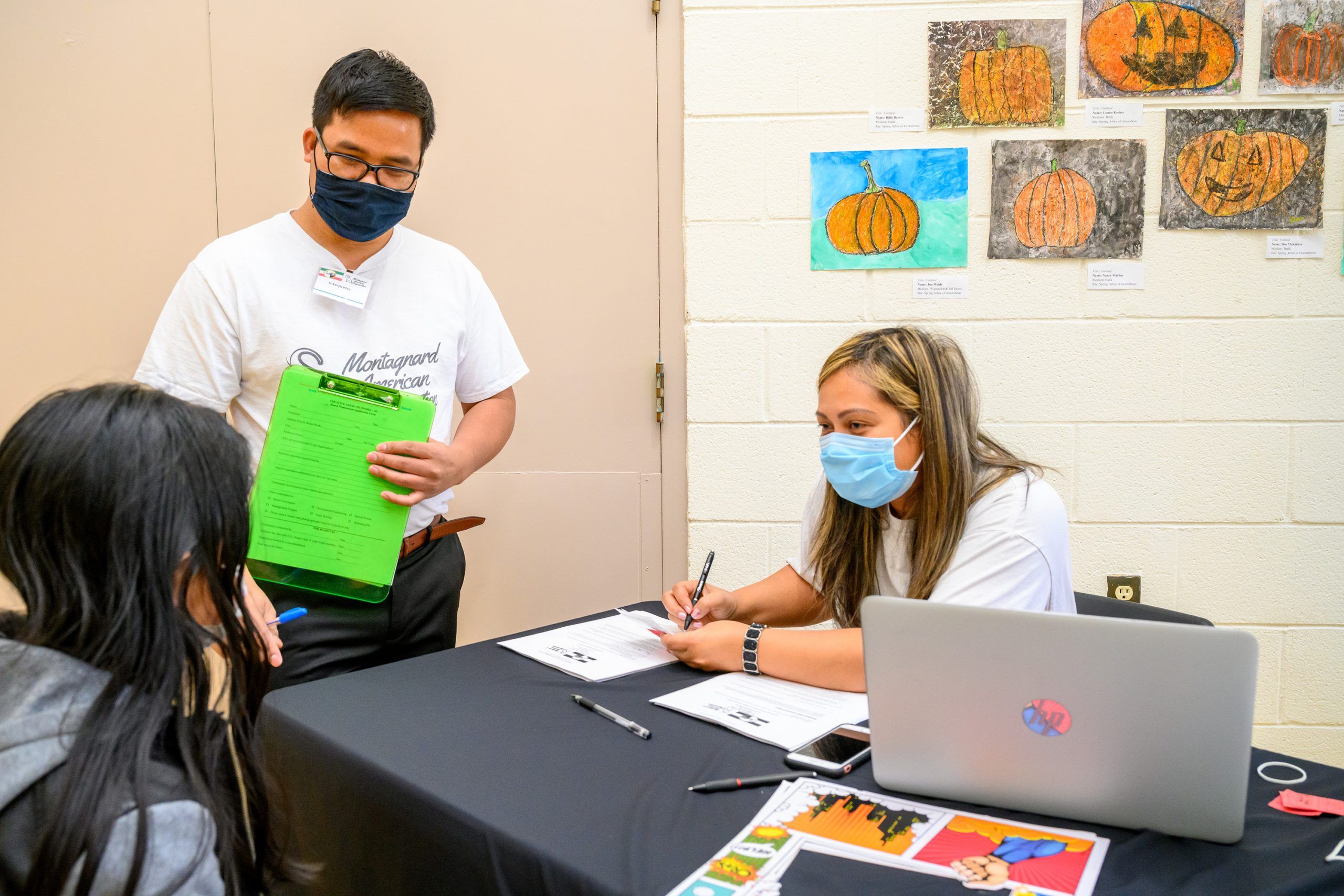 Siu standing next to a table, assisting community partner Liana H'Yua Adrong who is sittin at the table with intake at the vaccine clinic event.