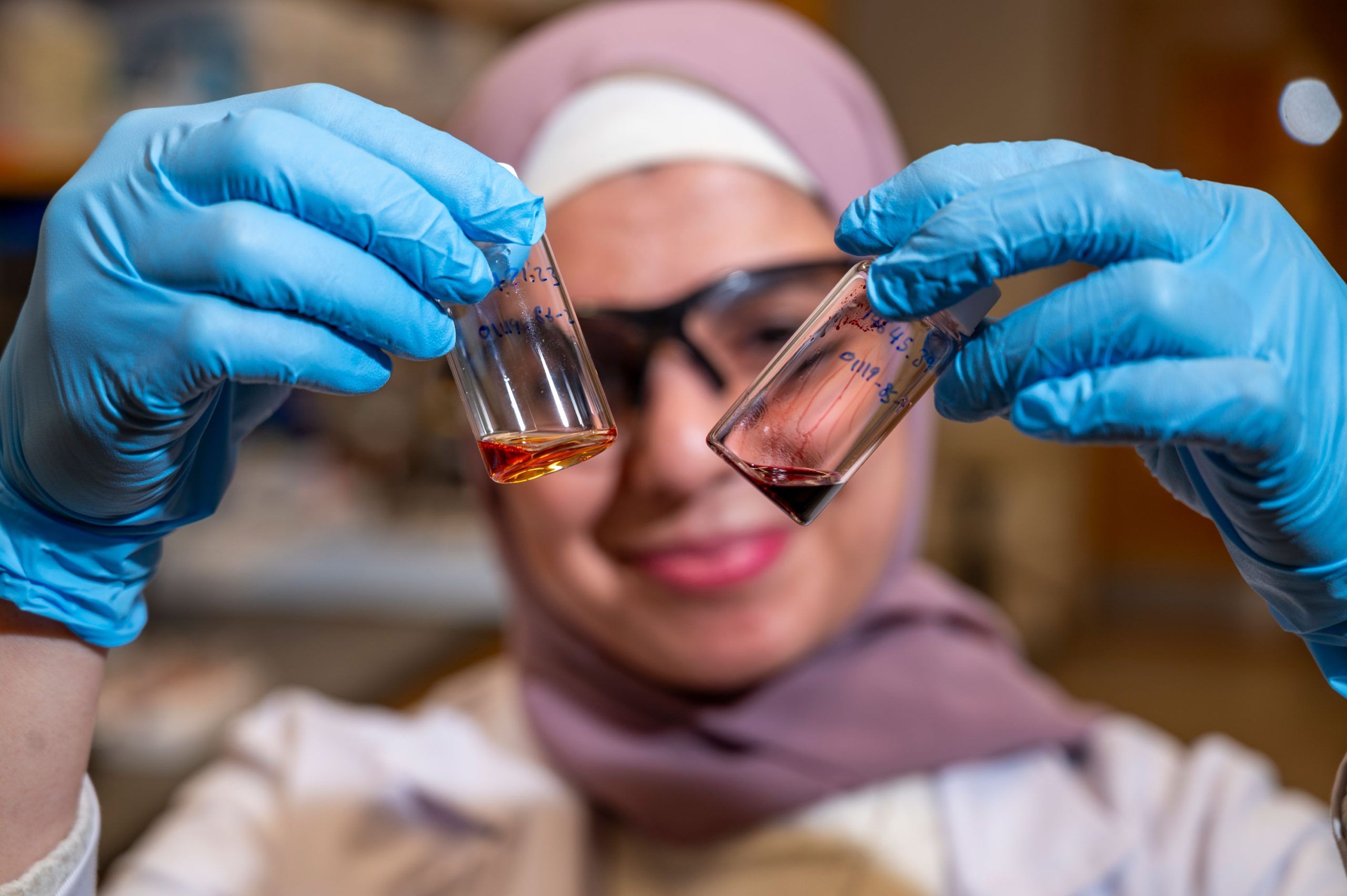 Dr. Al-Qiam's gloved hands holding a small clear vial with liquid in a laboratory setting. Shelves and equipment are visible in the blurred background.