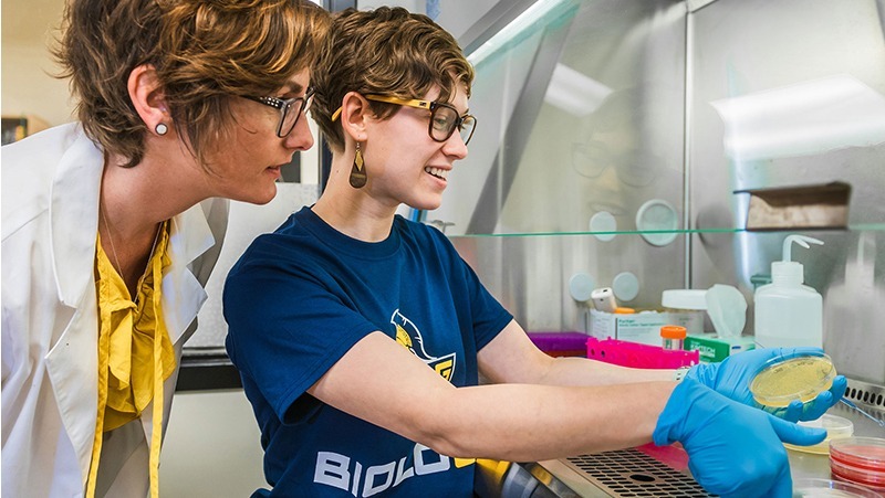 A person in a lab coat looks over the shoulder of a student holding a Petri dish
