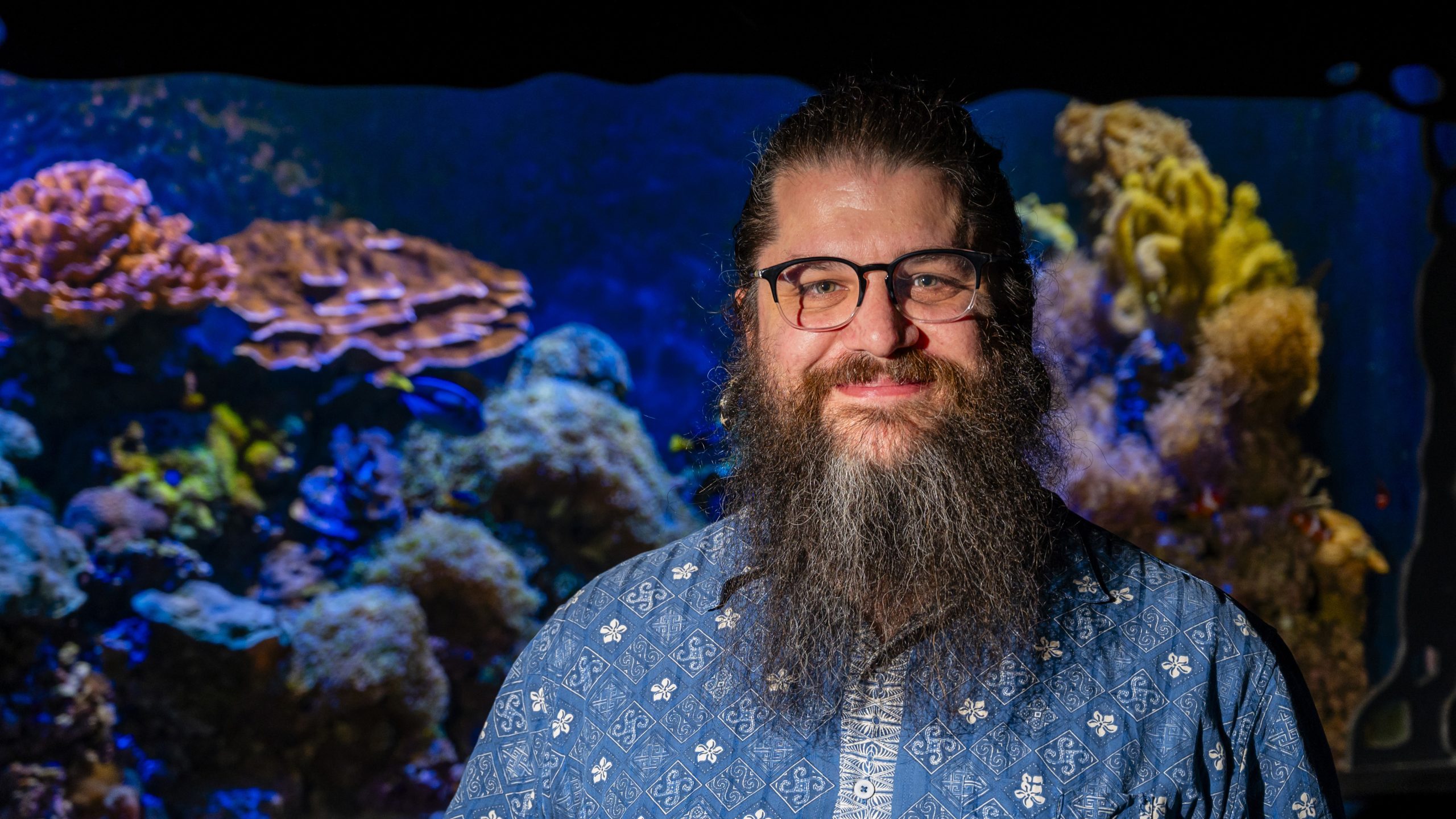 Dr. Derek Skillings smiles and stands in front of a coral reef tank
