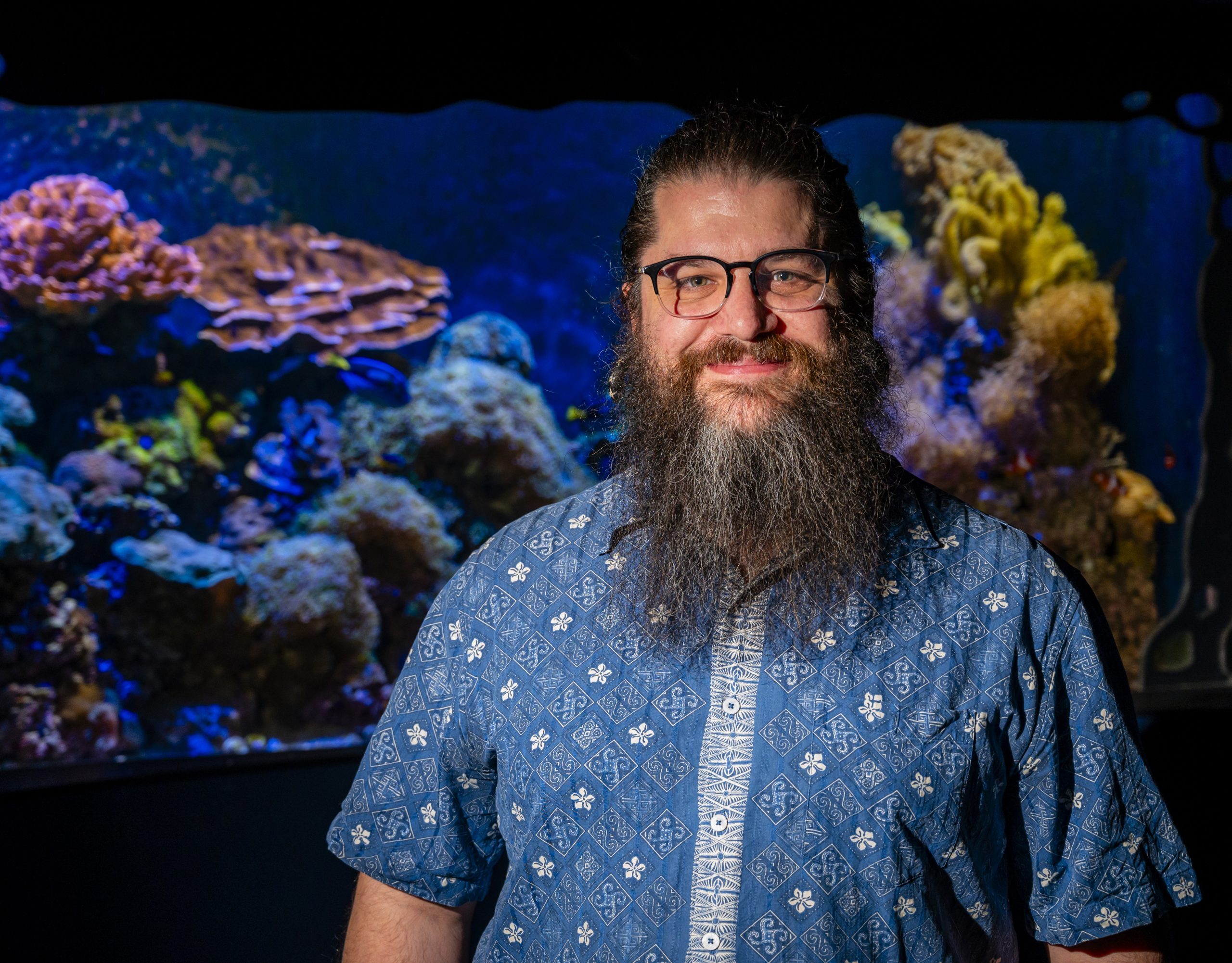 Dr. Derek Skilling stands and smiles in front of a coral reef tank