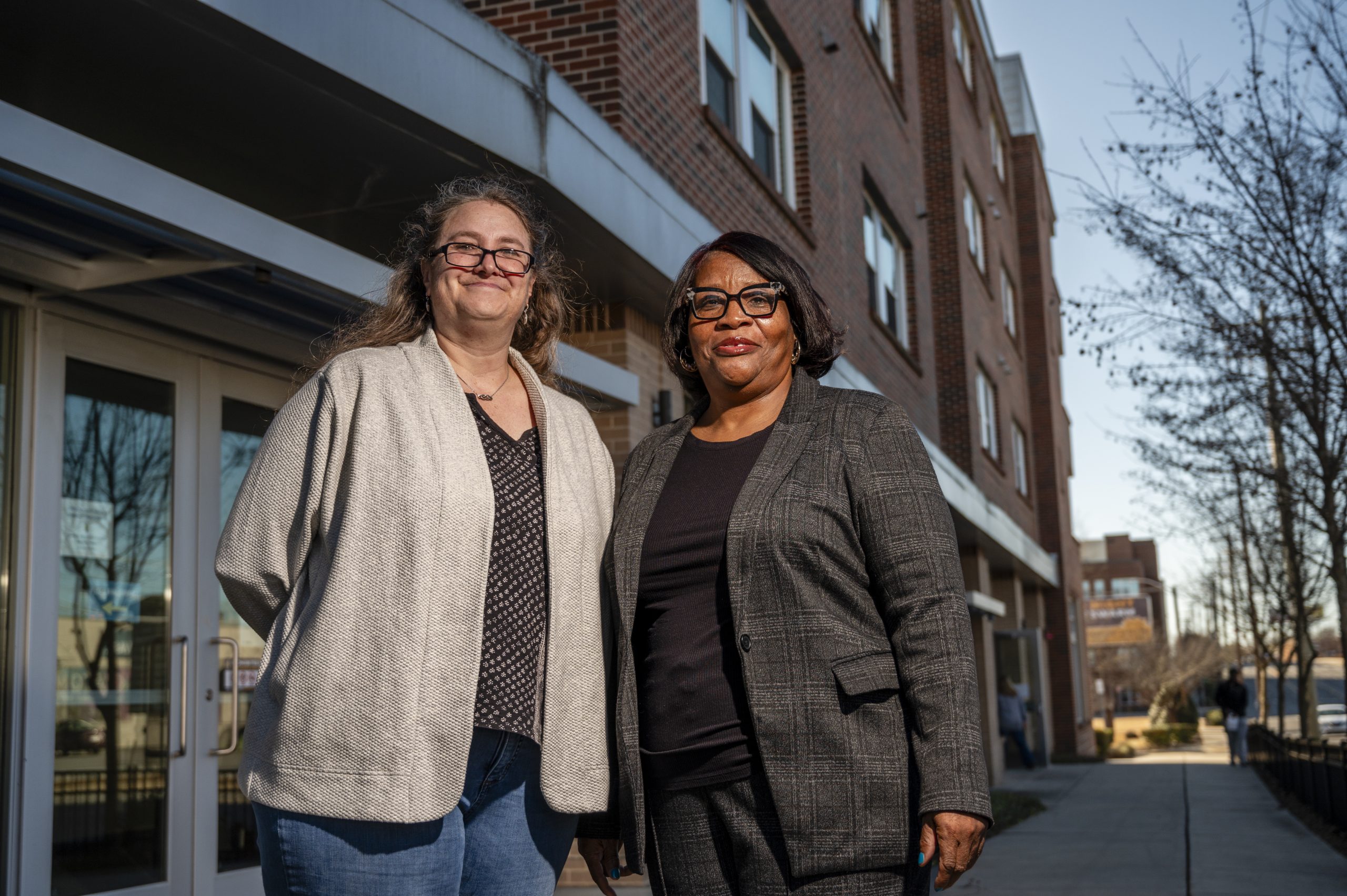 Willow Burgess-Johnson and Frederick Douglas standing outside a multi-story brick building with large windows and glass entrance doors. The sidewalk runs alongside the building, and leafless trees line the street under clear daylight.