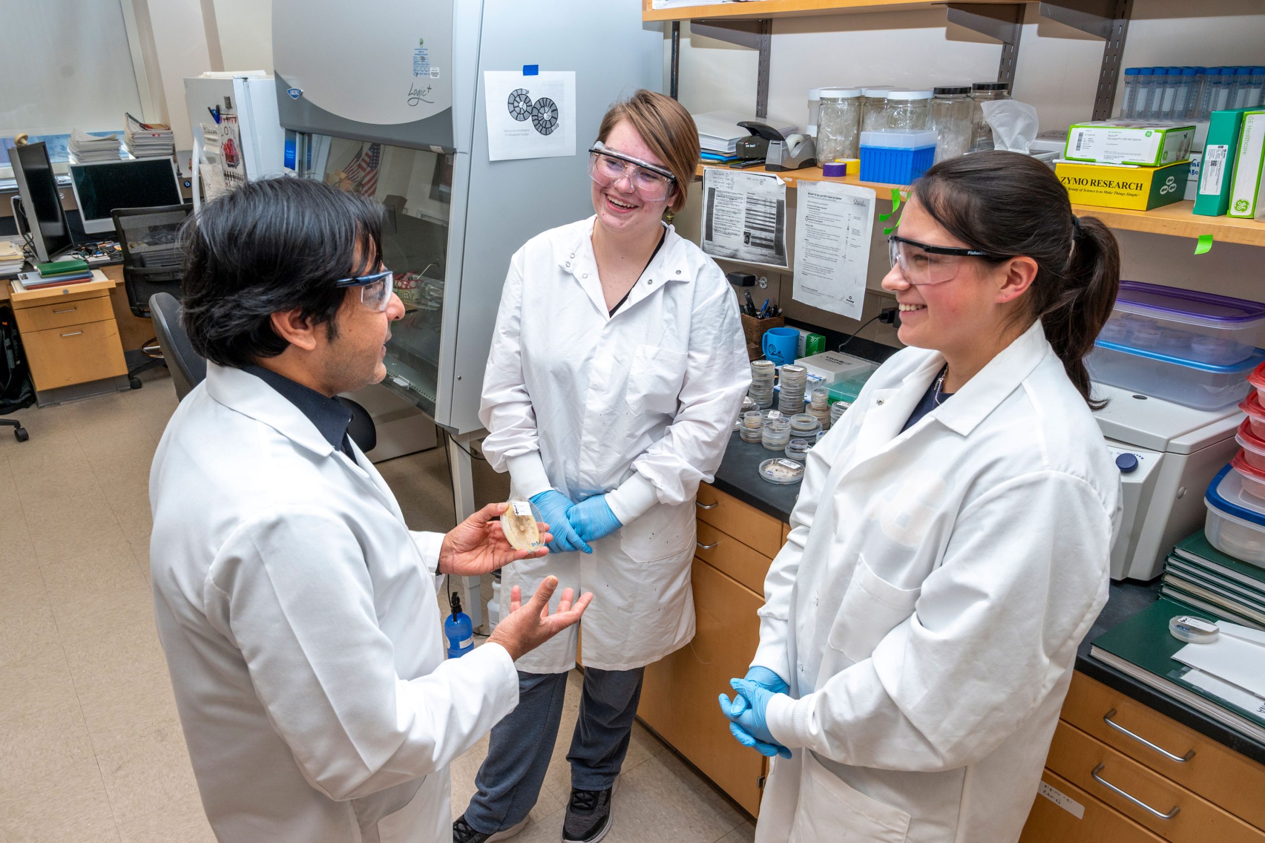 Dr. Raja and two students in a laboratory wearing white coats and gloves, standing near a counter with petri dishes and lab equipment. Dr. Raja is holding a sample while others listen. Background includes a fume hood, shelves with supplies, and printed diagrams on the wall.