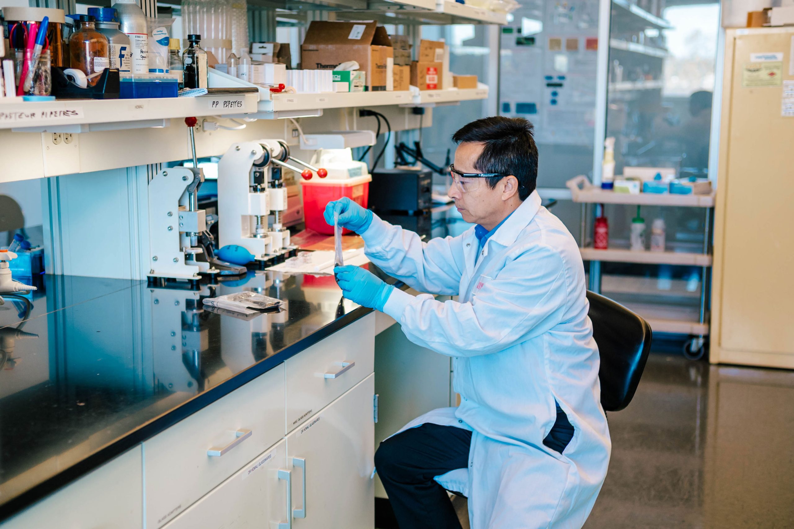 Dr. Wei in a laboratory wearing a white coat and blue gloves seated at a black countertop, holding a glass test tube with liquid. The workspace includes a microscope, pipettes, and various boxes and containers on shelves above.