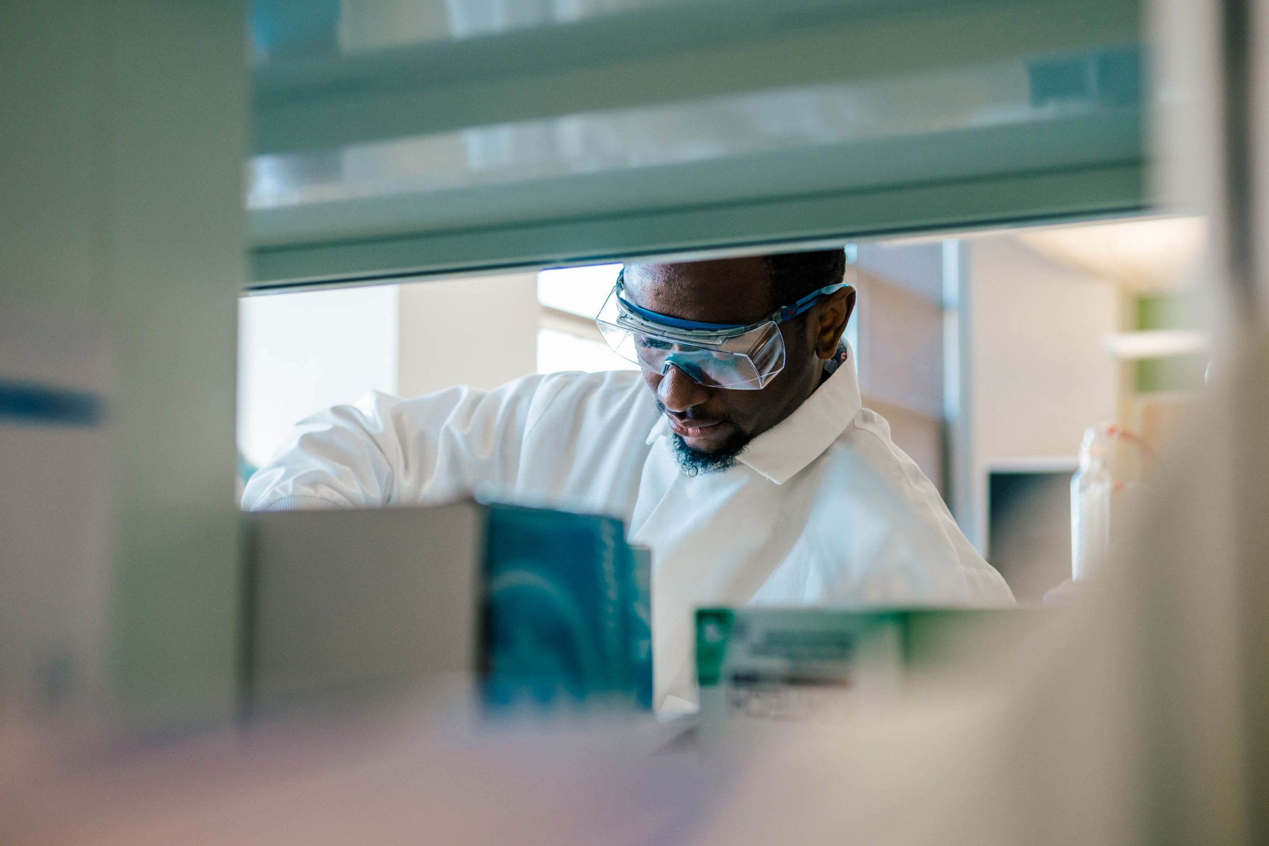 Laboratory view through a shelf opening showing a student in a white lab coat working among boxes and containers. Foreground includes packaged lab supplies and equipment partially visible on the counter.