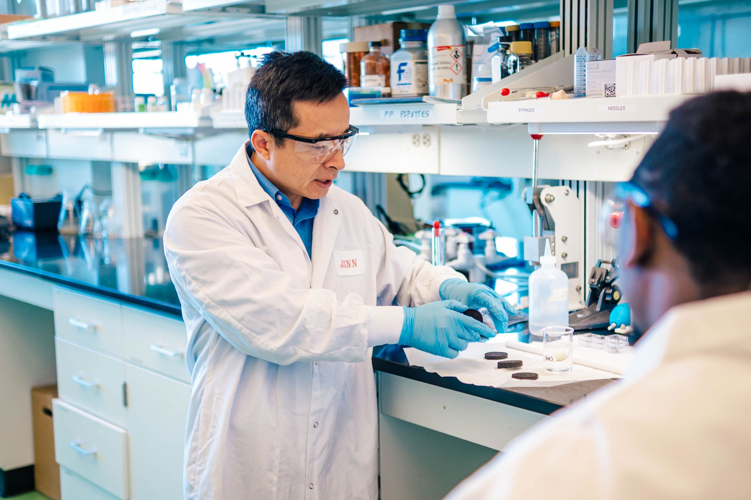 Dr. Wei in a laboratory wearing a white coat and blue gloves handling round black samples on a white surface. Nearby are a clear glass beaker, a bottle of liquid, and various lab supplies on a black countertop with shelves above holding containers and equipment.