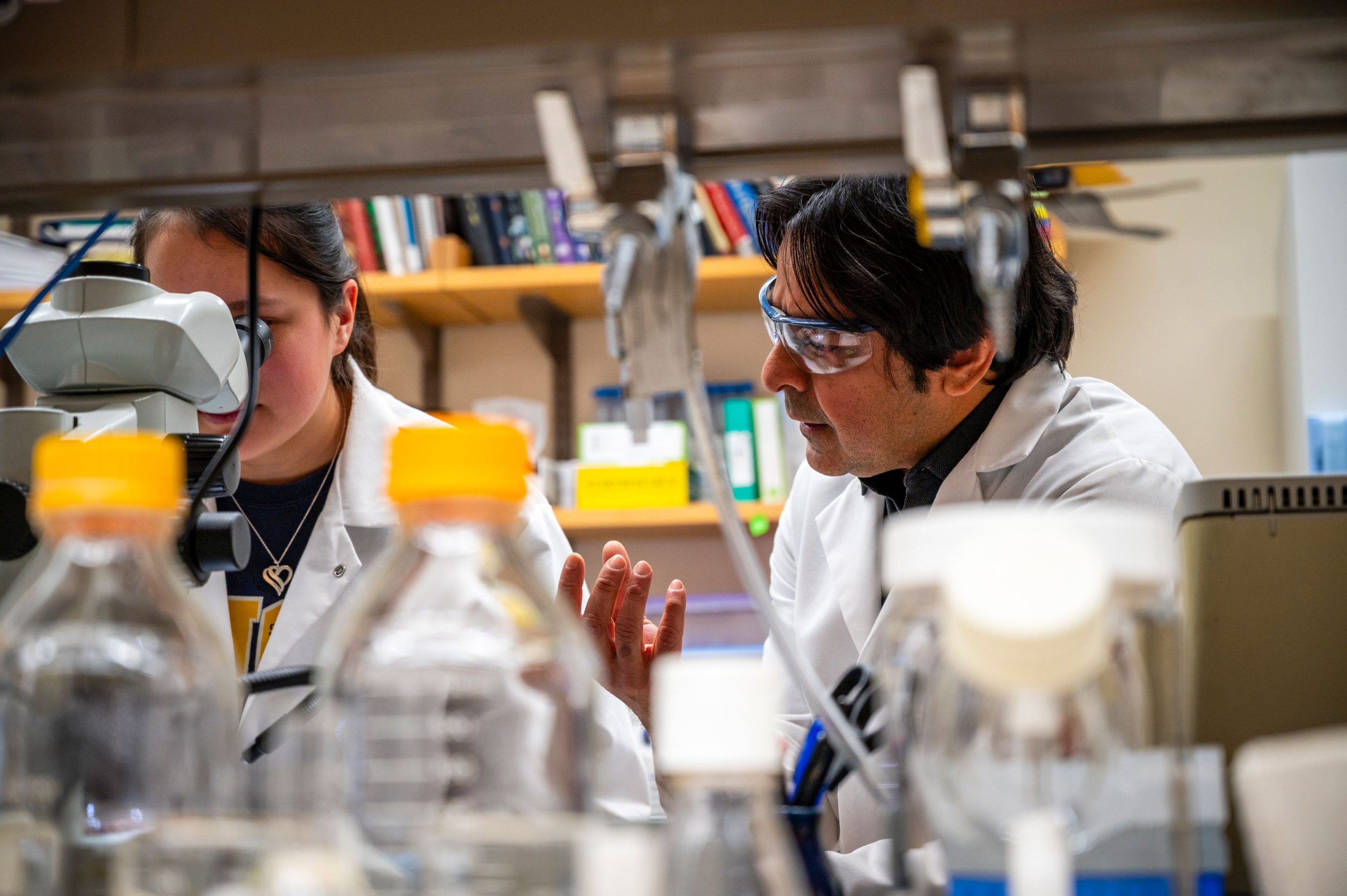 Laboratory scene showing Dr. Raja and a student in white coats seated at a bench surrounded by clear bottles with yellow caps, tubing, and other lab equipment. Shelves with books and supplies are visible in the background.