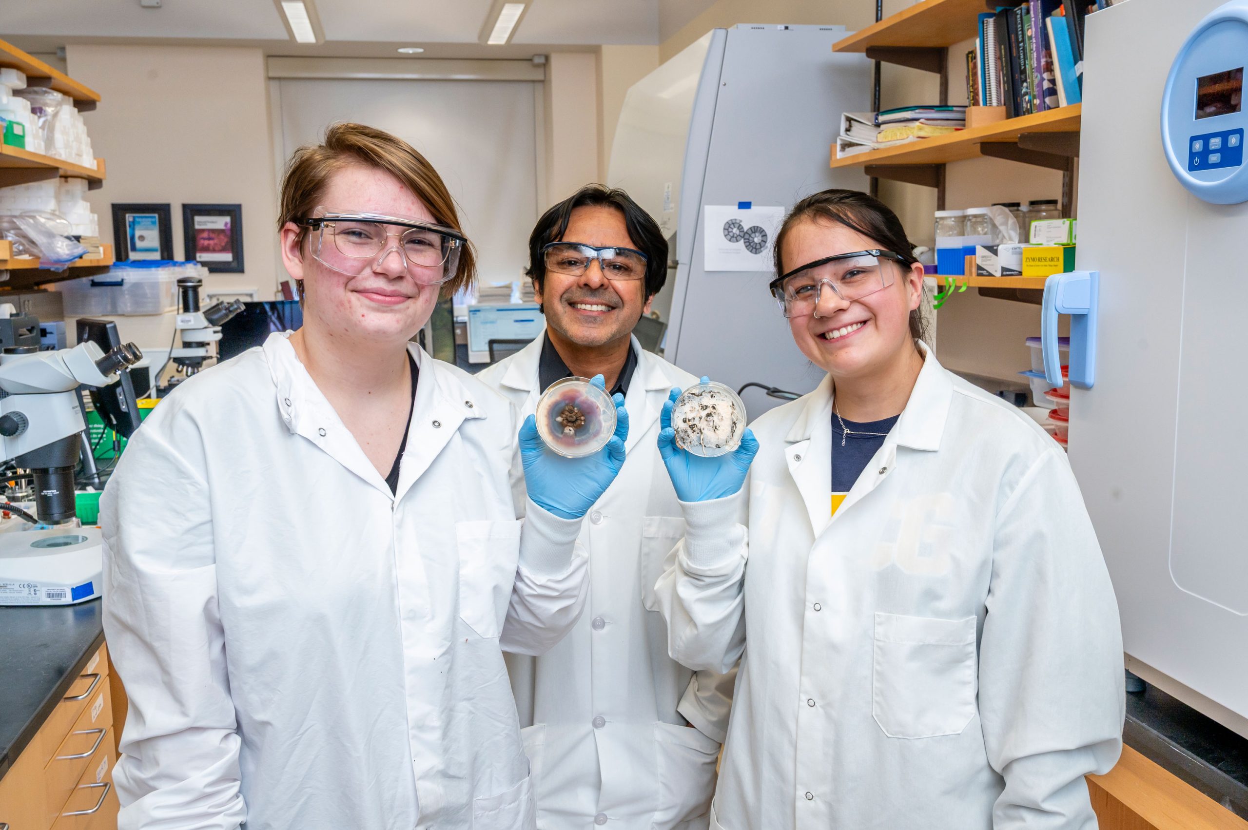 Dr. Raja and two of his students in a laboratory wearing white coats, with the two students holding petri dishes containing fungal cultures. Surrounding area includes shelves with books and supplies, a microscope, and lab equipment.