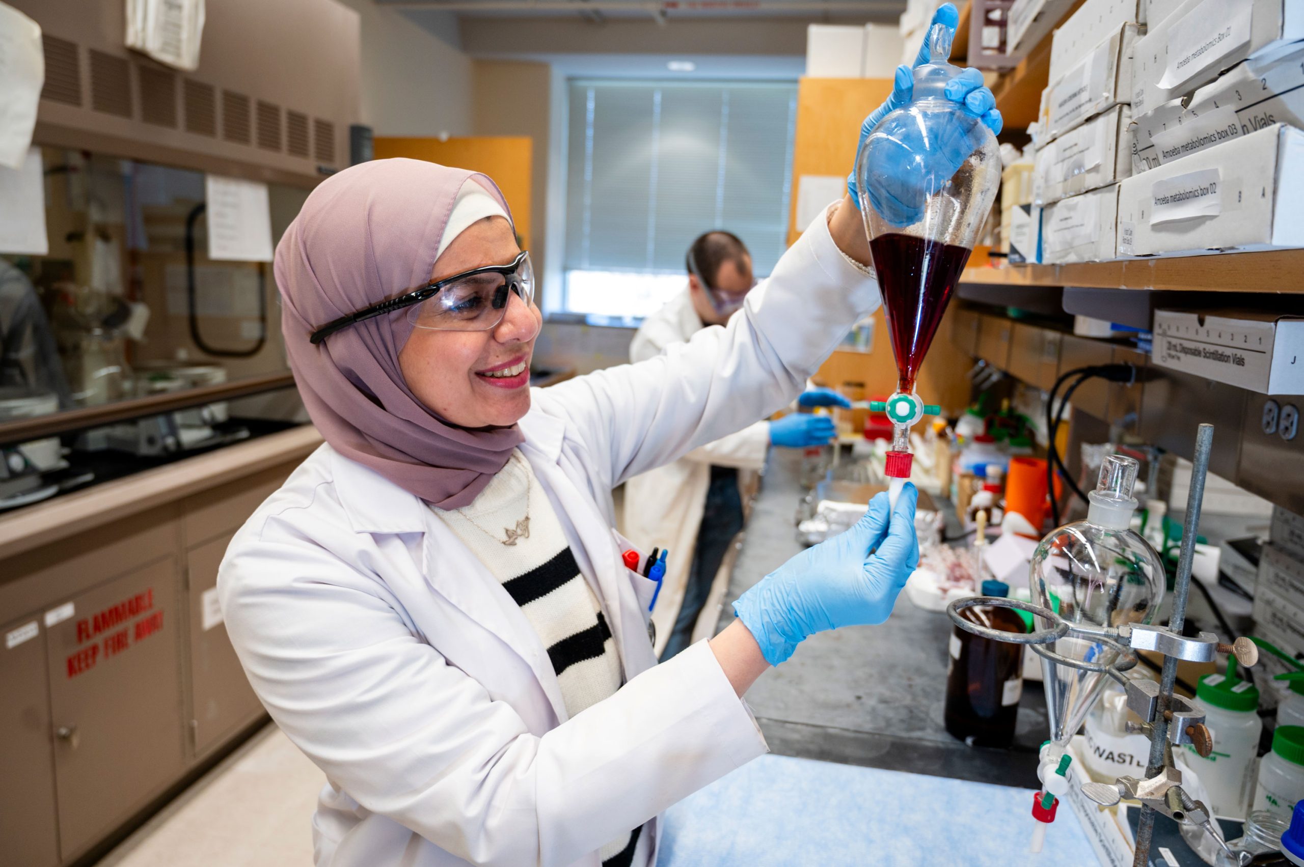 Laboratory scene showing Dr. Al-Qiam in a white coat and blue gloves holding a separatory funnel filled with dark red liquid, positioned over a clamp stand. The counter is lined with glassware, tubing, and labeled boxes, with additional lab equipment visible in the background.