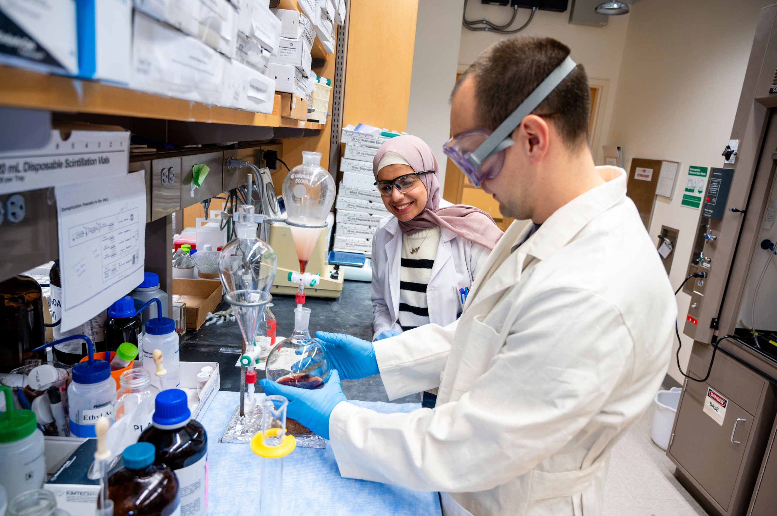 Laboratory scene showing Al-Qiam and a student working at a counter with a separatory funnel containing dark liquid held in a clamp stand. The bench is lined with glassware, bottles, and labeled boxes, with additional lab equipment visible in the background.