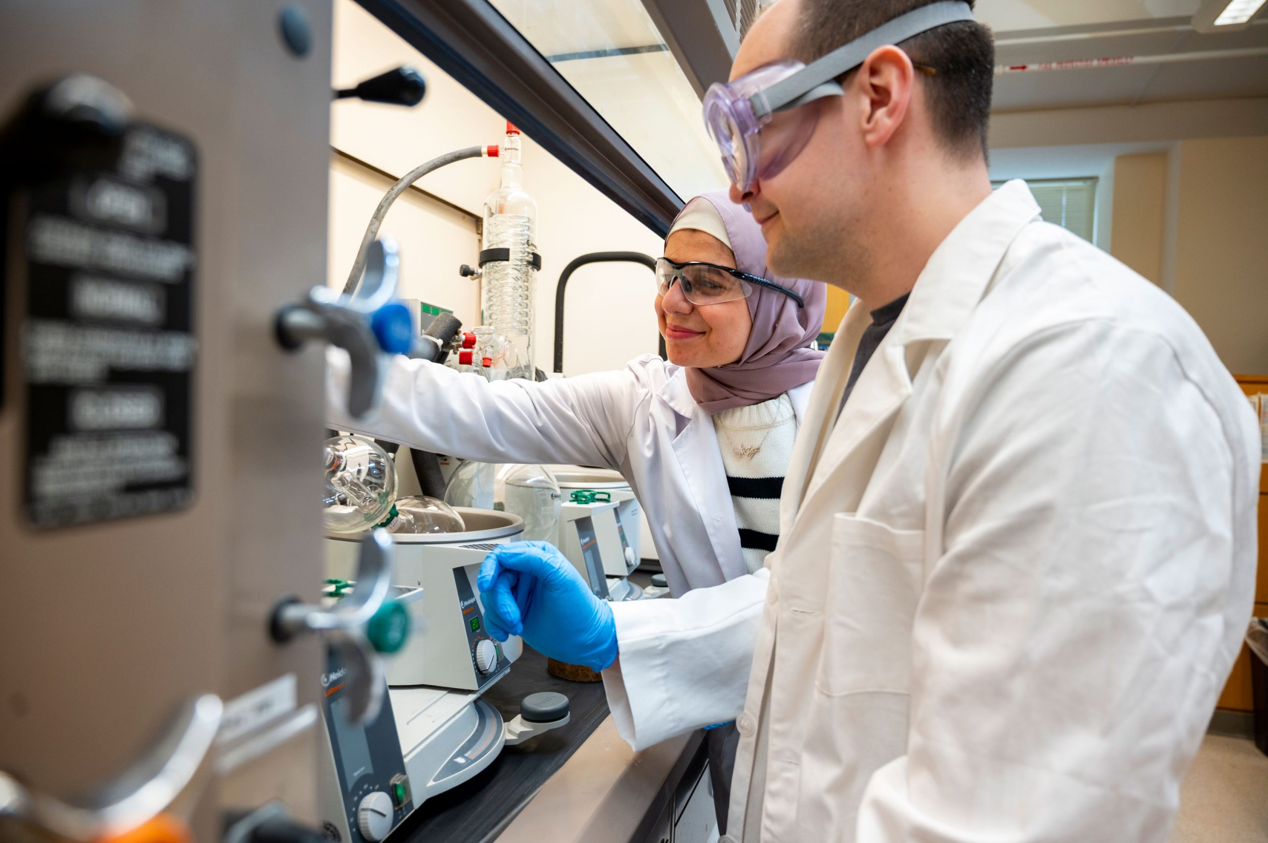 Al-Qiam and another student in a laboratory working with a rotary evaporator inside a fume hood. One person adjusts the control knob while the other reaches toward the glass apparatus connected to tubing and a condenser column. Various lab equipment and clamps are visible on the bench.