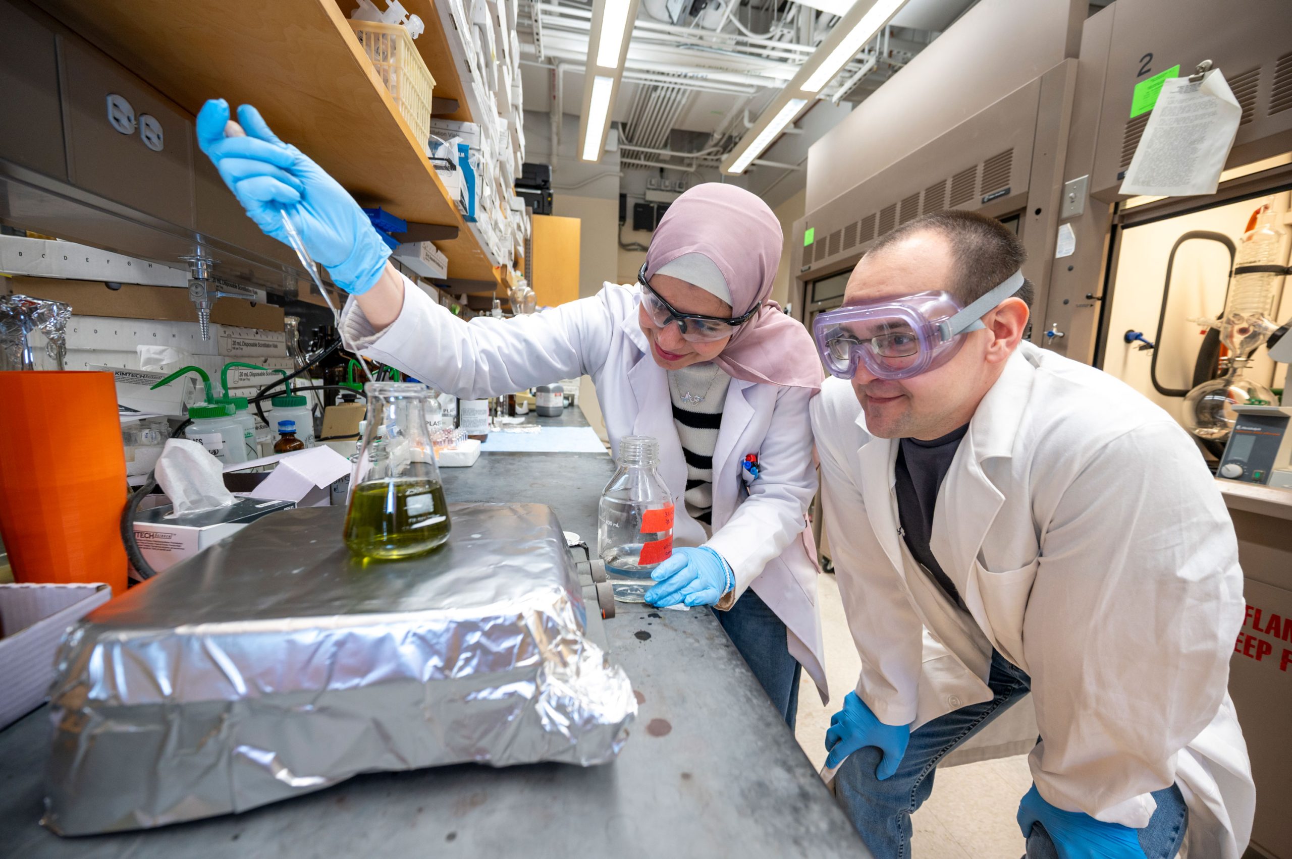Al-Qiam and another student in a laboratory working at a counter with glass flasks containing green liquid. One person is adjusting a flask on a metal surface covered with foil, while the other observes. Shelves above hold various containers and lab supplies.