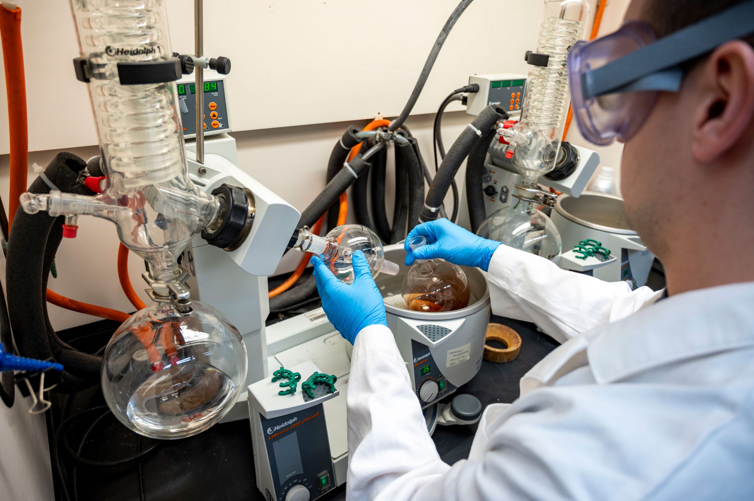 Laboratory setup showing a person wearing gloves operating a rotary evaporator. The apparatus includes glass condenser coils, round-bottom flasks, and tubing connected to a heating bath partially filled with liquid.