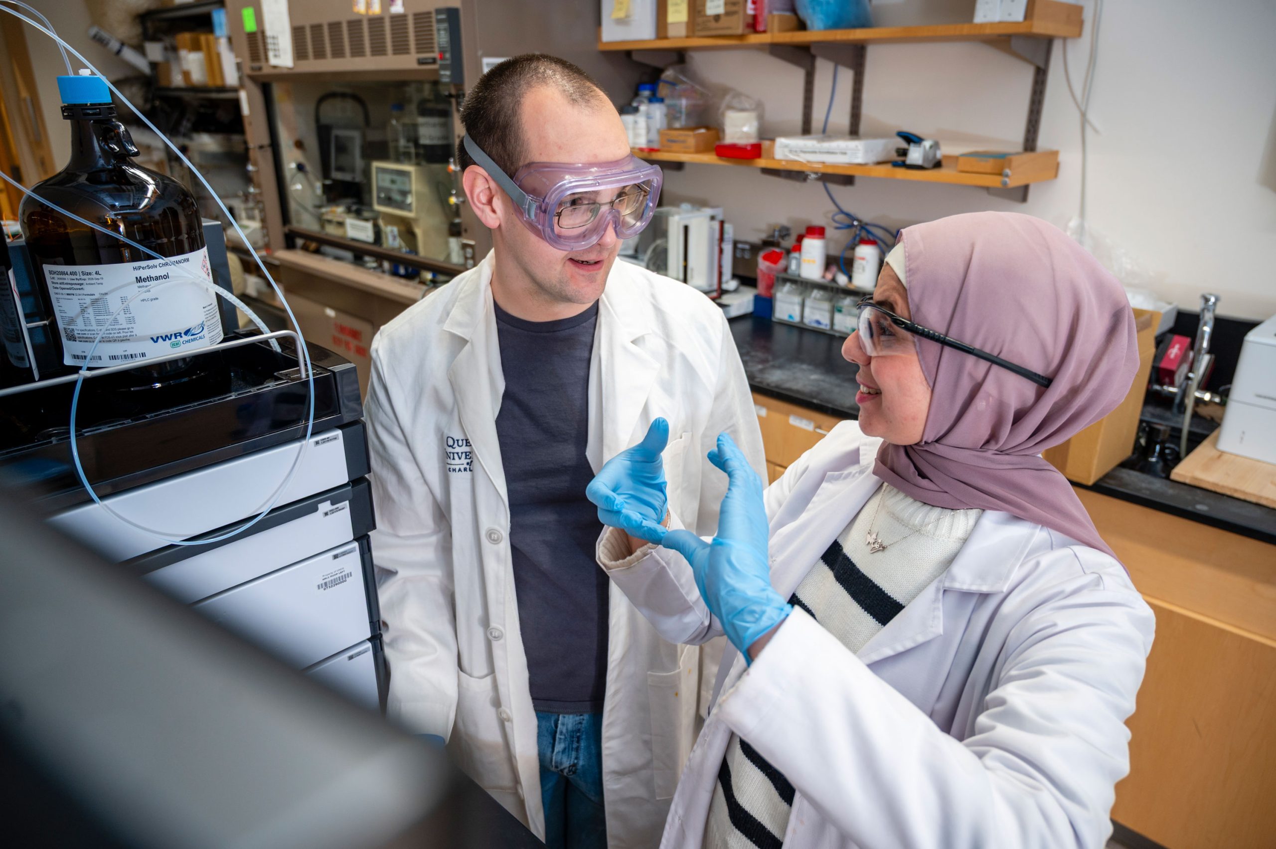 Al-Qiam and a student in a laboratory standing near a chromatography system with tubing and large reagent bottles. Al-Qiam is gesturing with gloved hands while the student observes. Shelves above hold various containers and equipment.