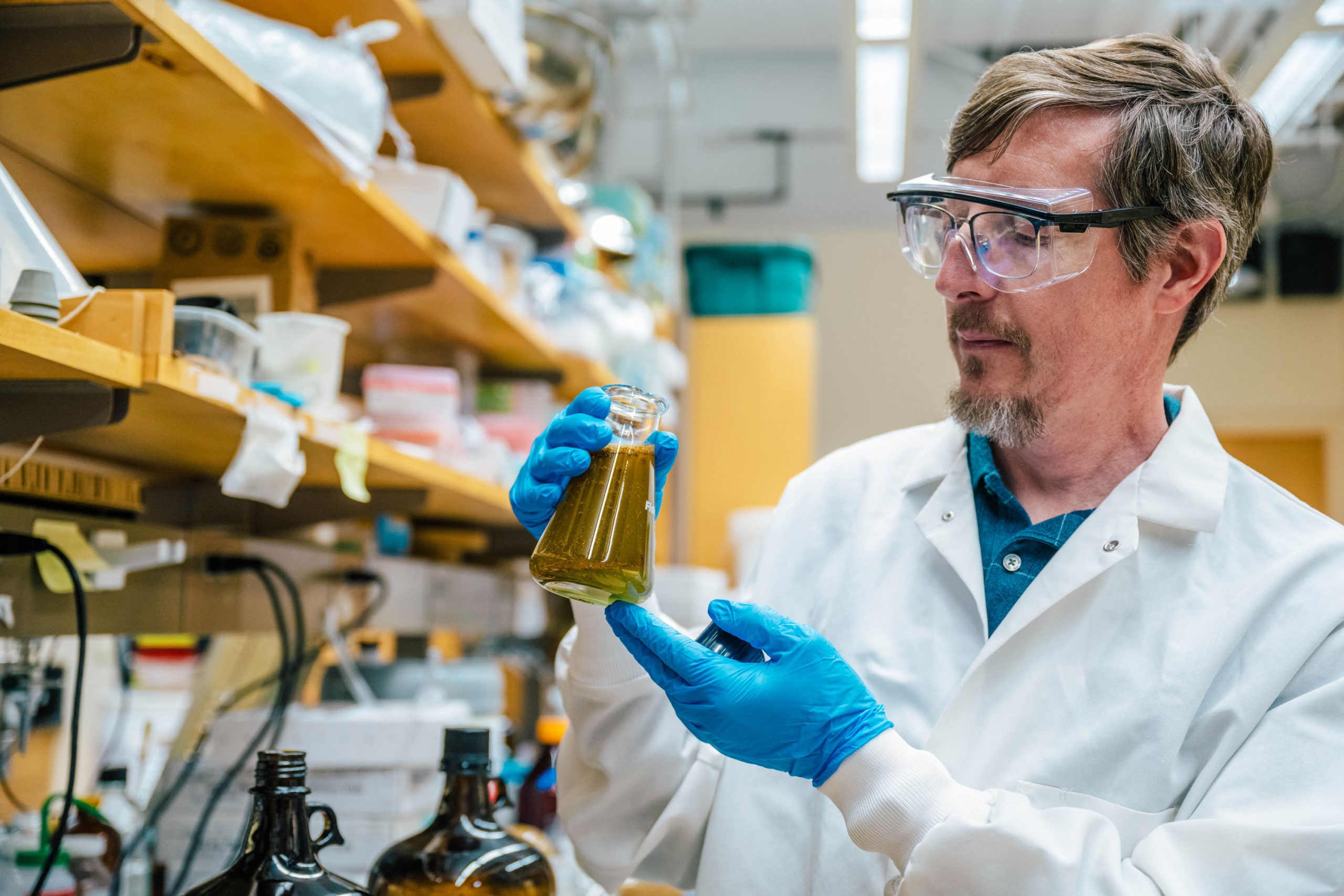 Tyler Graf wearing a white lab coat and blue gloves holding a glass flask filled with greenish liquid in a laboratory. Shelves with bottles, containers, and equipment are visible in the background.