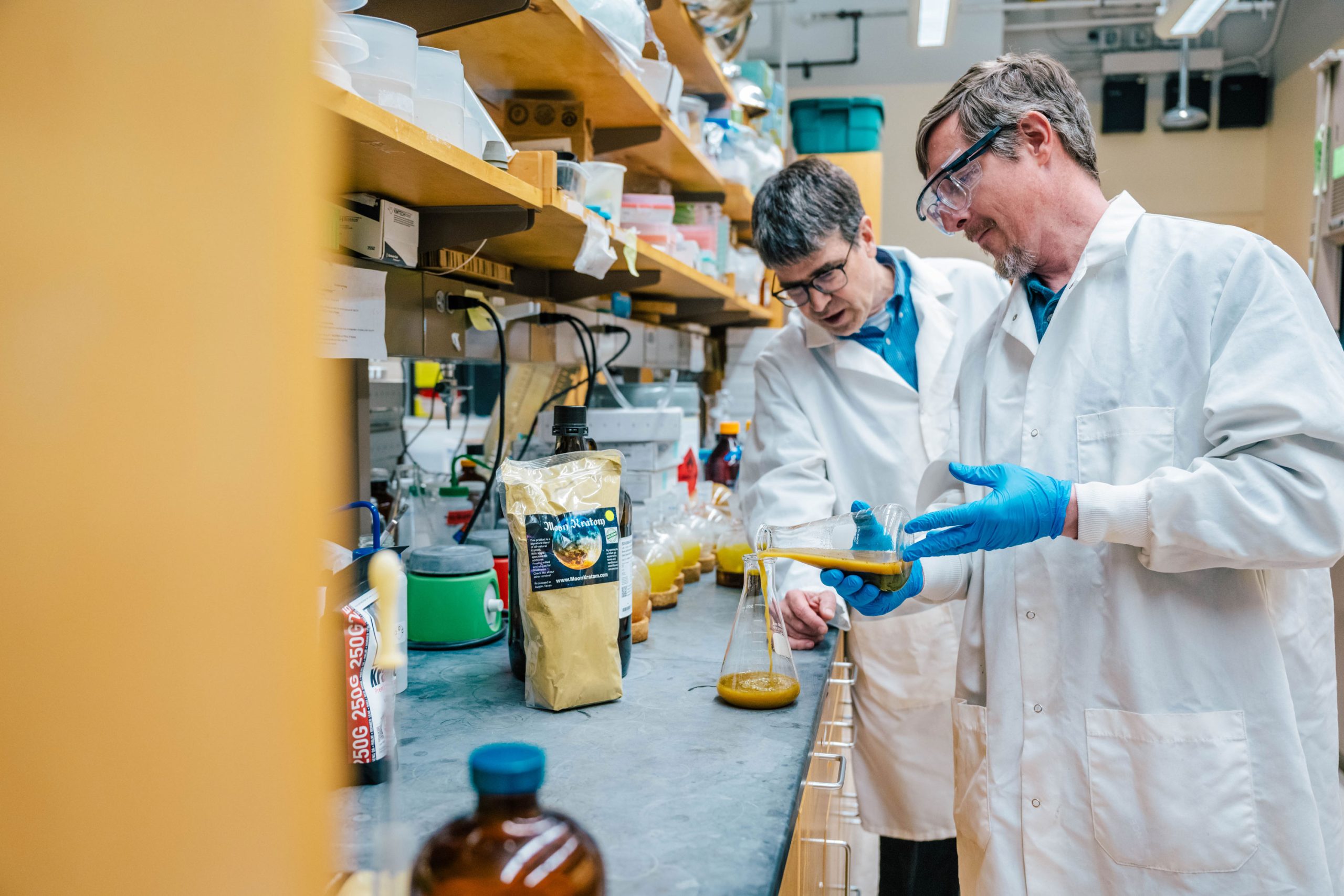 Tyler Graf and Dr. Oberlies in a laboratory wearing white coats and blue gloves working at a counter. Graf is pouring a thick yellow liquid from a graduated cylinder into a glass flask. The counter holds various containers, including a large pouch labeled “Organic Protein,” bottles, and lab equipment.
