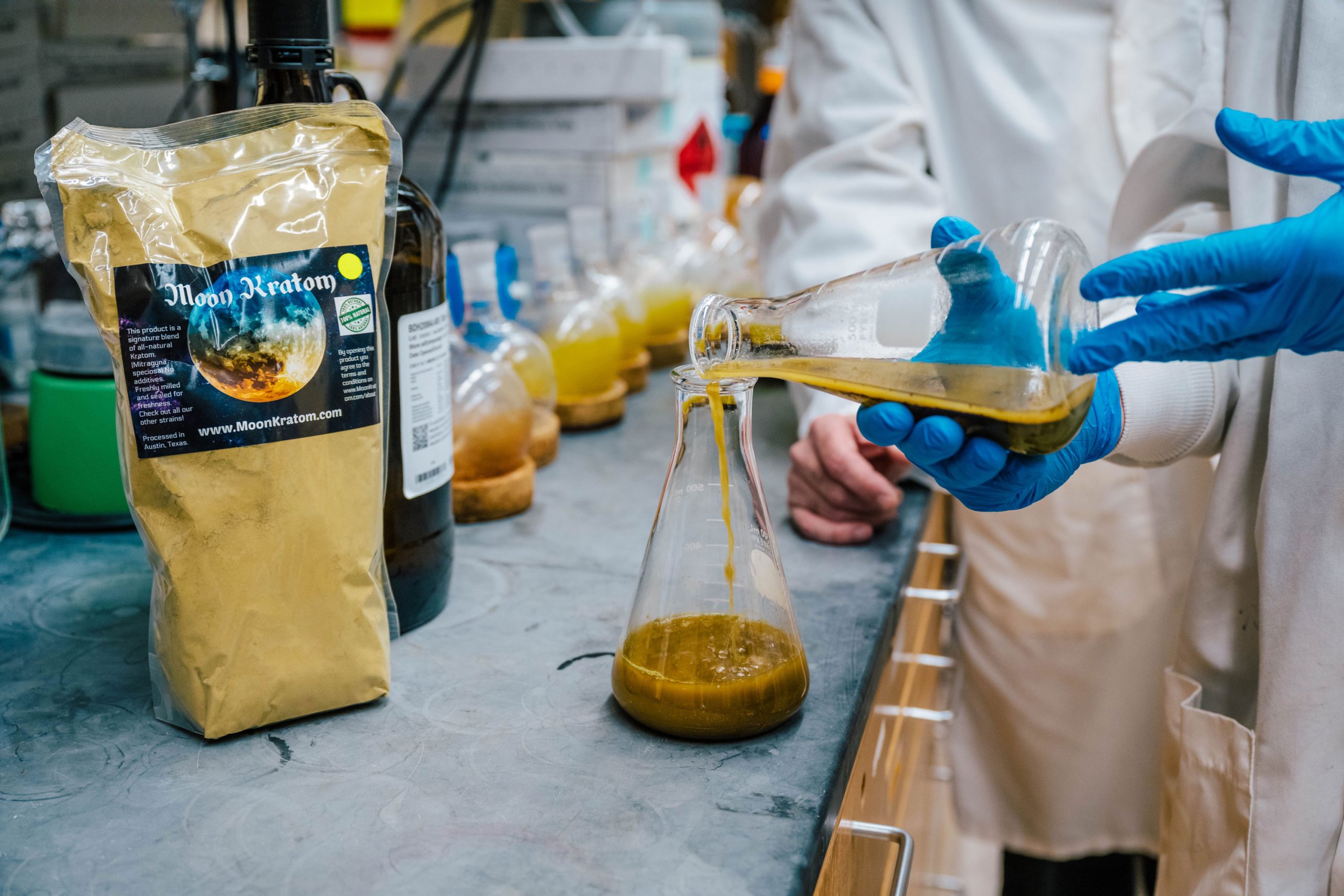 Gloved hands pouring a thick greenish-brown liquid from one glass flask into another on a lab bench. Nearby are additional flasks, a bag labeled “Moon Kratom,” and other laboratory supplies.