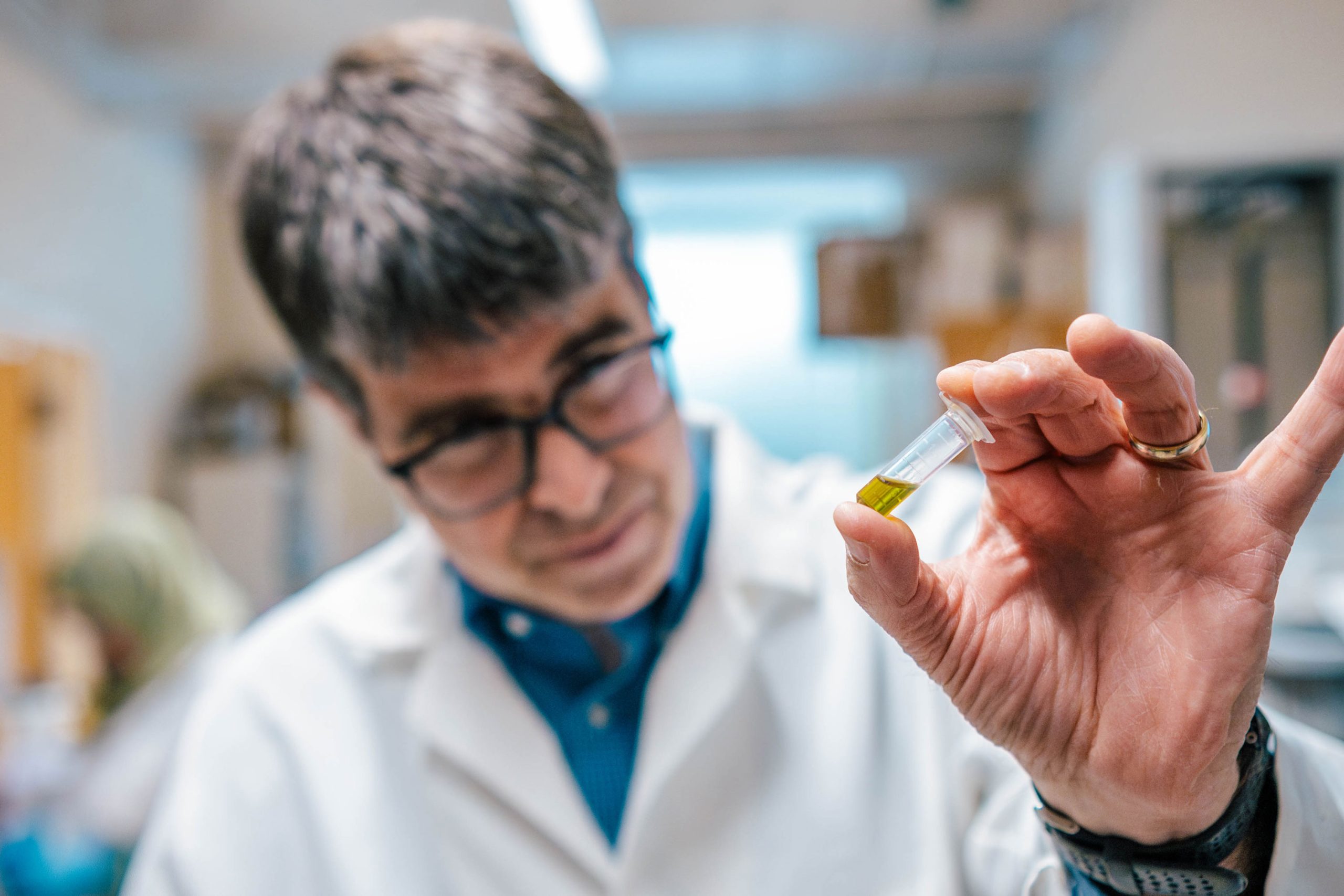 Close-up of Dr. Oberlies' hand holding a small clear vial containing yellow liquid in a laboratory setting. Background shows shelves and equipment slightly out of focus.