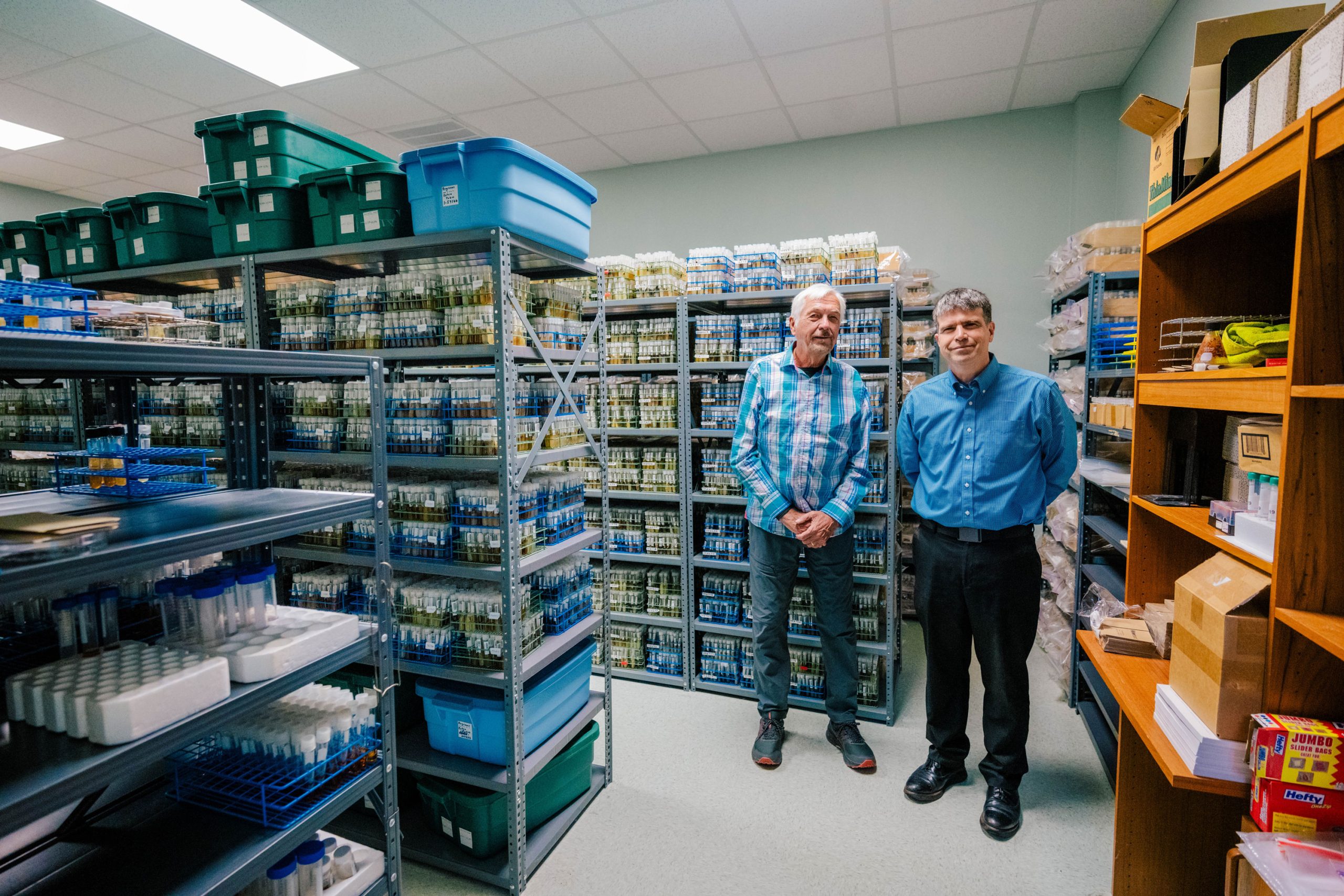 Cedric Pearce and Dr. Oberlies standing in a storage room filled with metal shelving units holding hundreds of labeled test tubes in blue racks, along with plastic bins and boxes.
