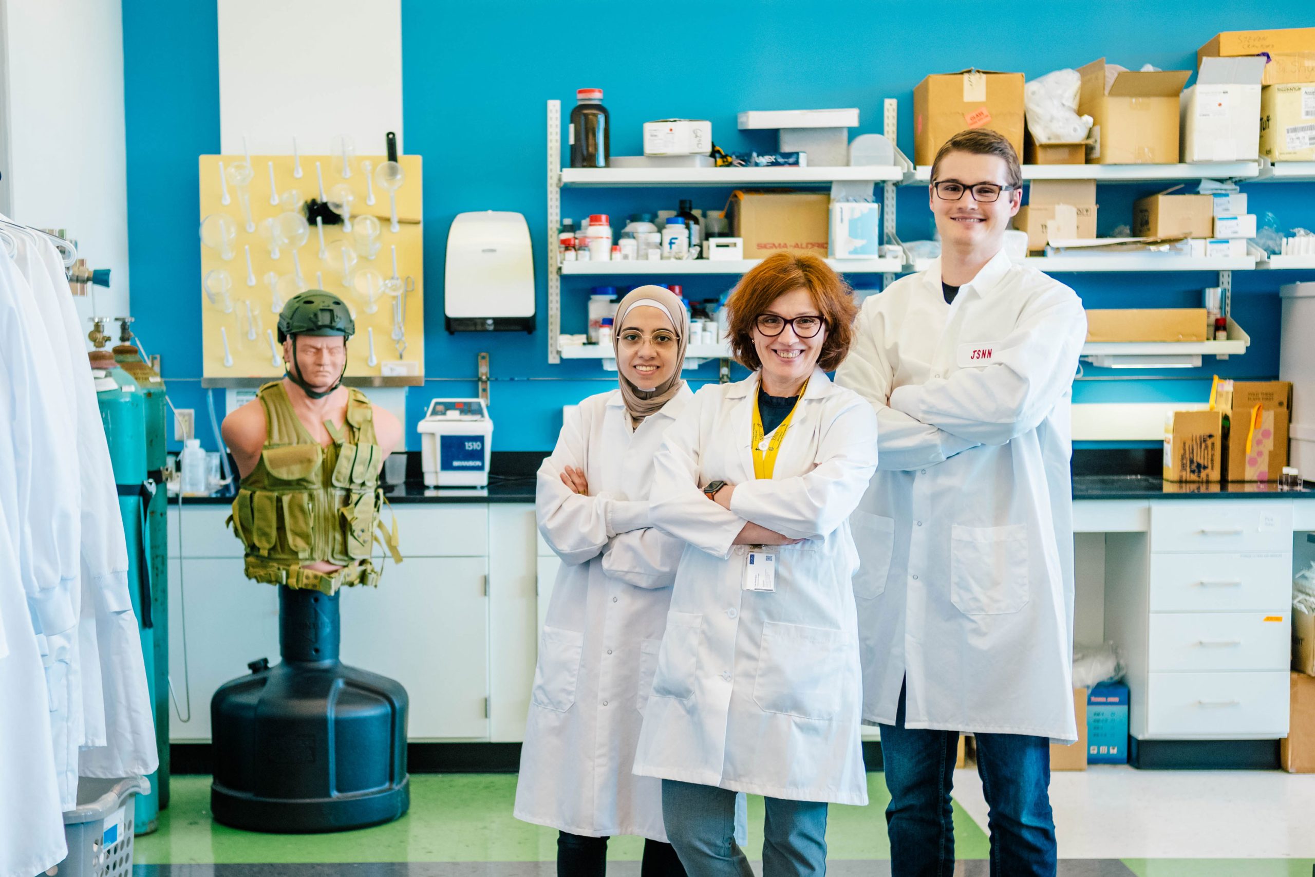Dr. Ignatova and two of her students wearing white lab coats standing in a laboratory with arms crossed. Behind them are shelves filled with boxes and equipment, a blue wall-mounted paper towel dispenser, and a mannequin torso wearing a green tactical vest. The workspace includes various lab tools and supplies.